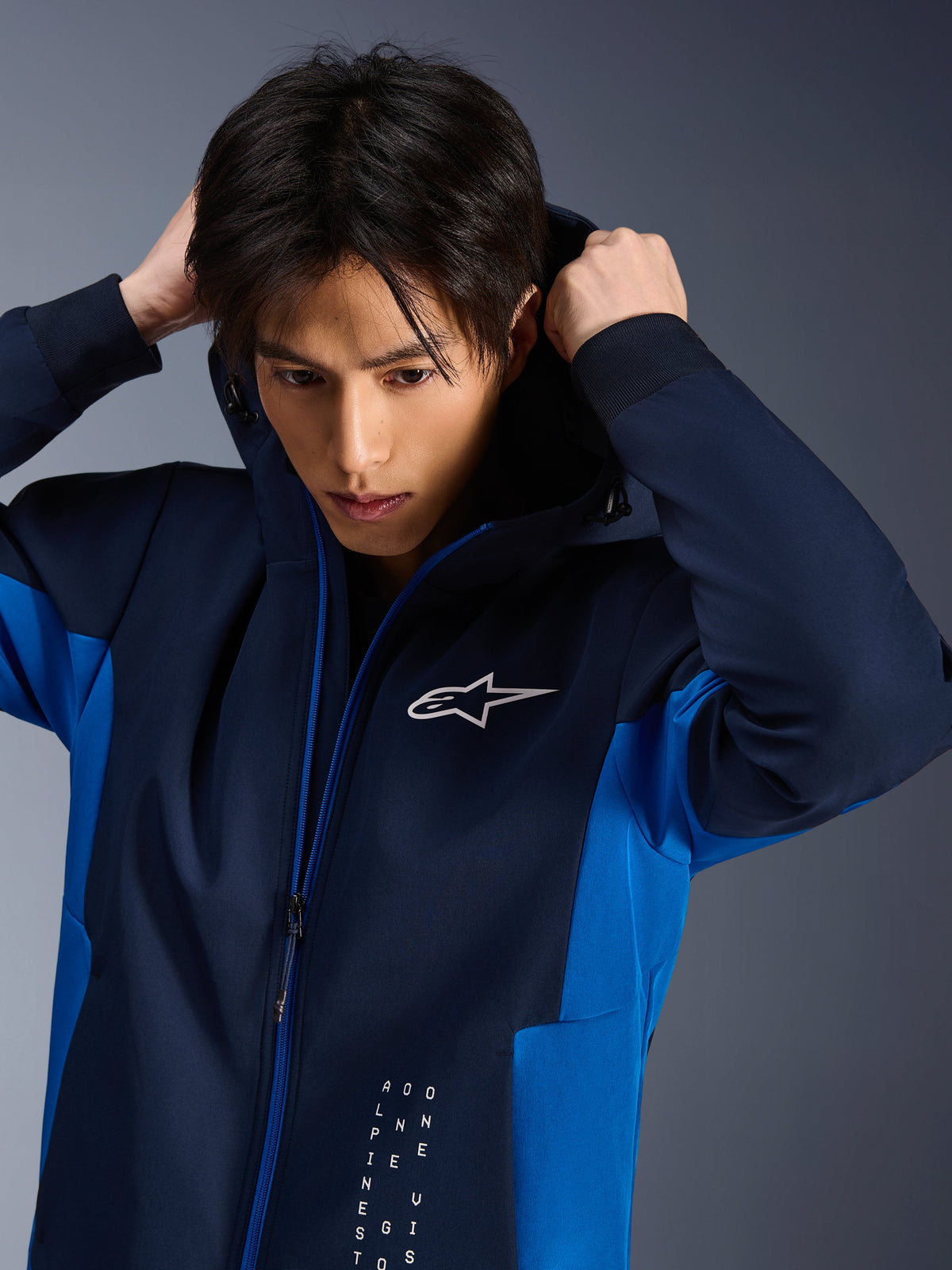 Alpinestars Radium Tech Hoodie, giacca da moto urbana, Night Navy e Royal Blue, indossata da un modello che si sistema il cappuccio, con logo Astars bianco sul petto e guscio tecnico 3L con vestibilità atletica.