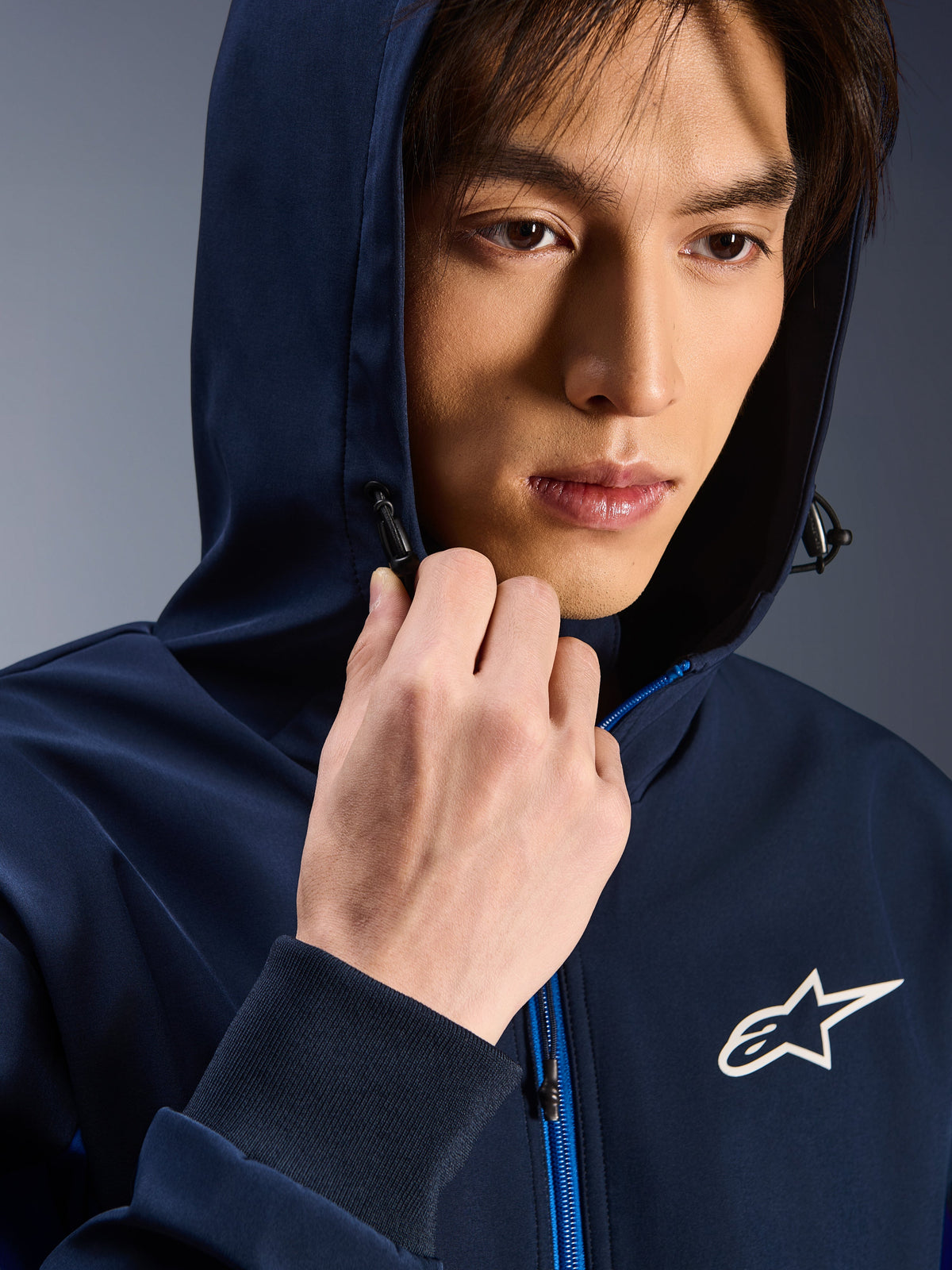 Alpinestars Radium Tech Hoodie, Night Navy Royal Blue, primo piano di un uomo che indossa la felpa con cappuccio alzato, logo Astars bianco sul petto, cerniera blu, guscio tecnico da moto urbano