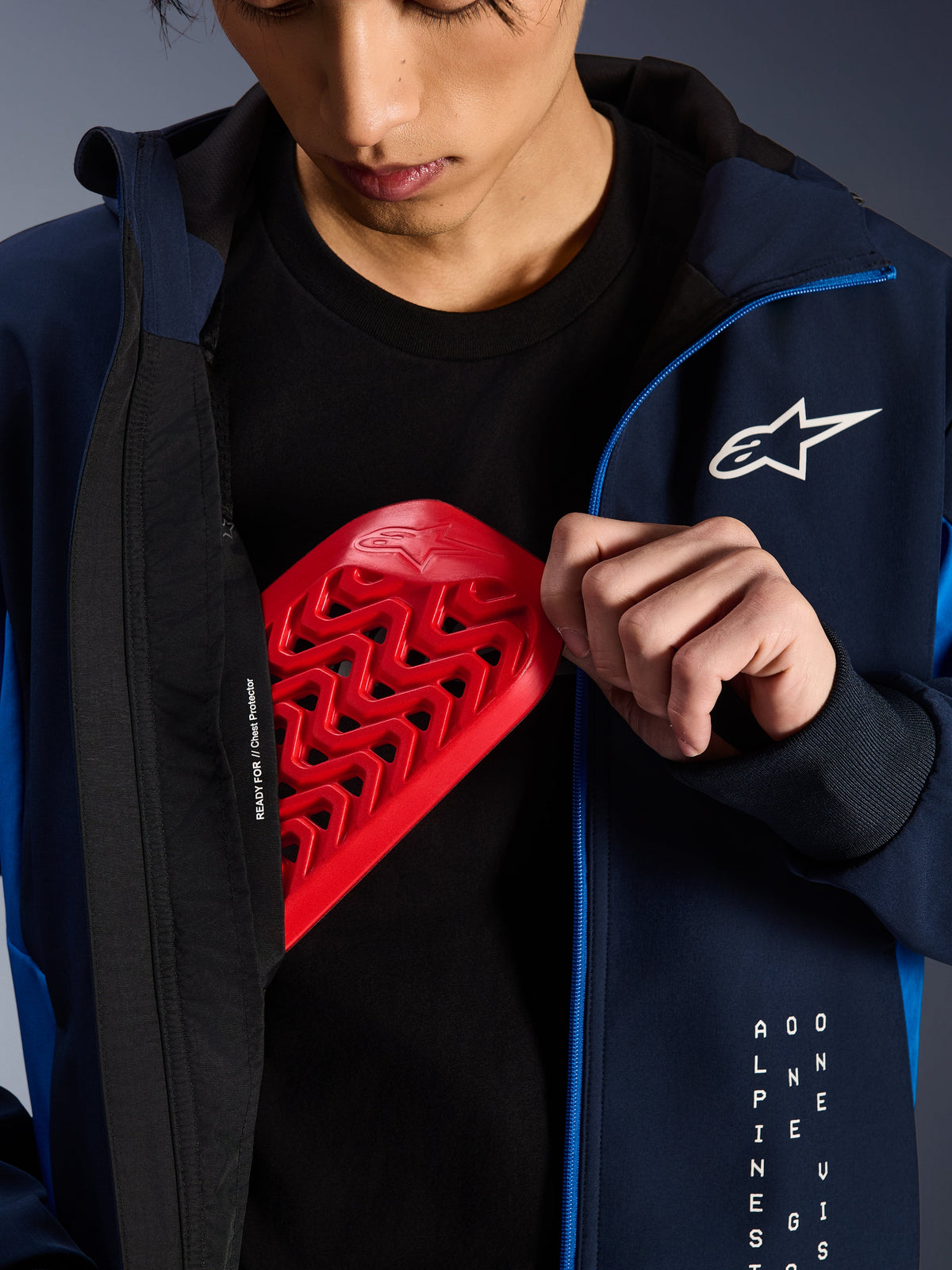 Alpinestars Radium Tech Hoodie nei colori Night Navy e Royal Blue, primo piano che mostra l'inserimento di una protezione toracica Nucleon rossa nella tasca interna, con la scritta 'READY FOR // Chest Protector' sulla fodera e un logo a stella bianco sul petto.