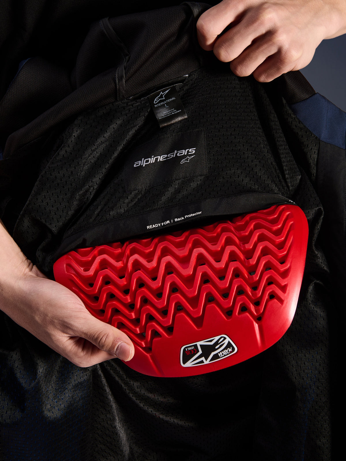 Dettaglio interno della felpa Alpinestars Radium Tech Hoodie, paraschiena rosso Nucleon Plasma inserito nella tasca dedicata, fodera in mesh nera con scritta 'READY FOR | Back Protector', design a nido d'ape ondulato, caratteristiche protettive per giacca da moto urbana tecnica