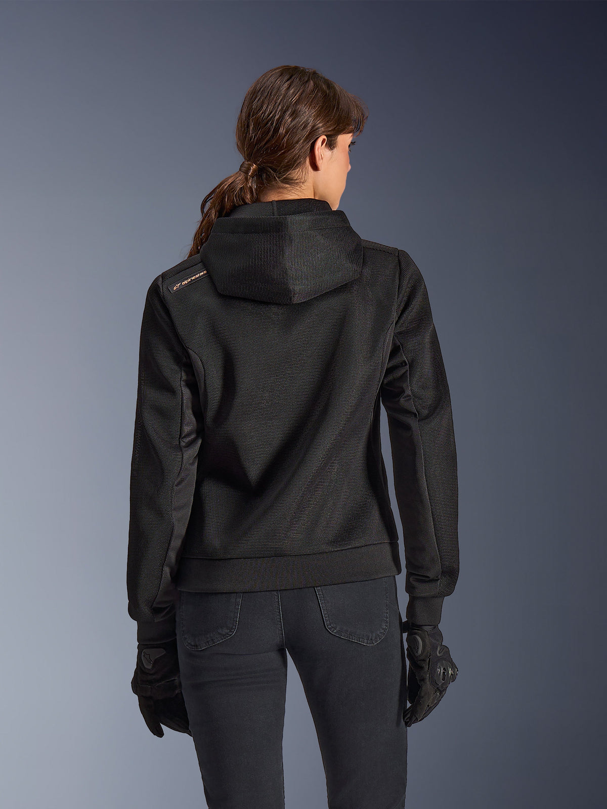 Alpinestars Stella Chrome Superairflow Hoodie, vista posteriore di donna con felpa tecnica da moto in mesh 3D nera, cappuccio ingegnerizzato e logo color rame sulla spalla, indossata con guanti e pantaloni da moto neri.