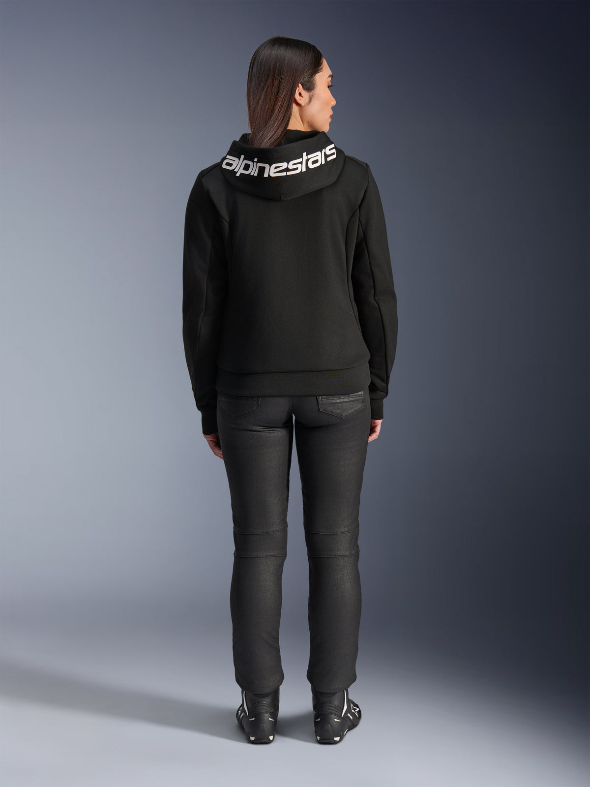 Vista posteriore di una modella che indossa la felpa Alpinestars Stella Chrome V2 Sport Hoodie nera, con un evidente logo Alpinestars bianco sul cappuccio, abbinata a pantaloni da moto neri e stivali da motociclista, abbigliamento da moto urban sport da donna