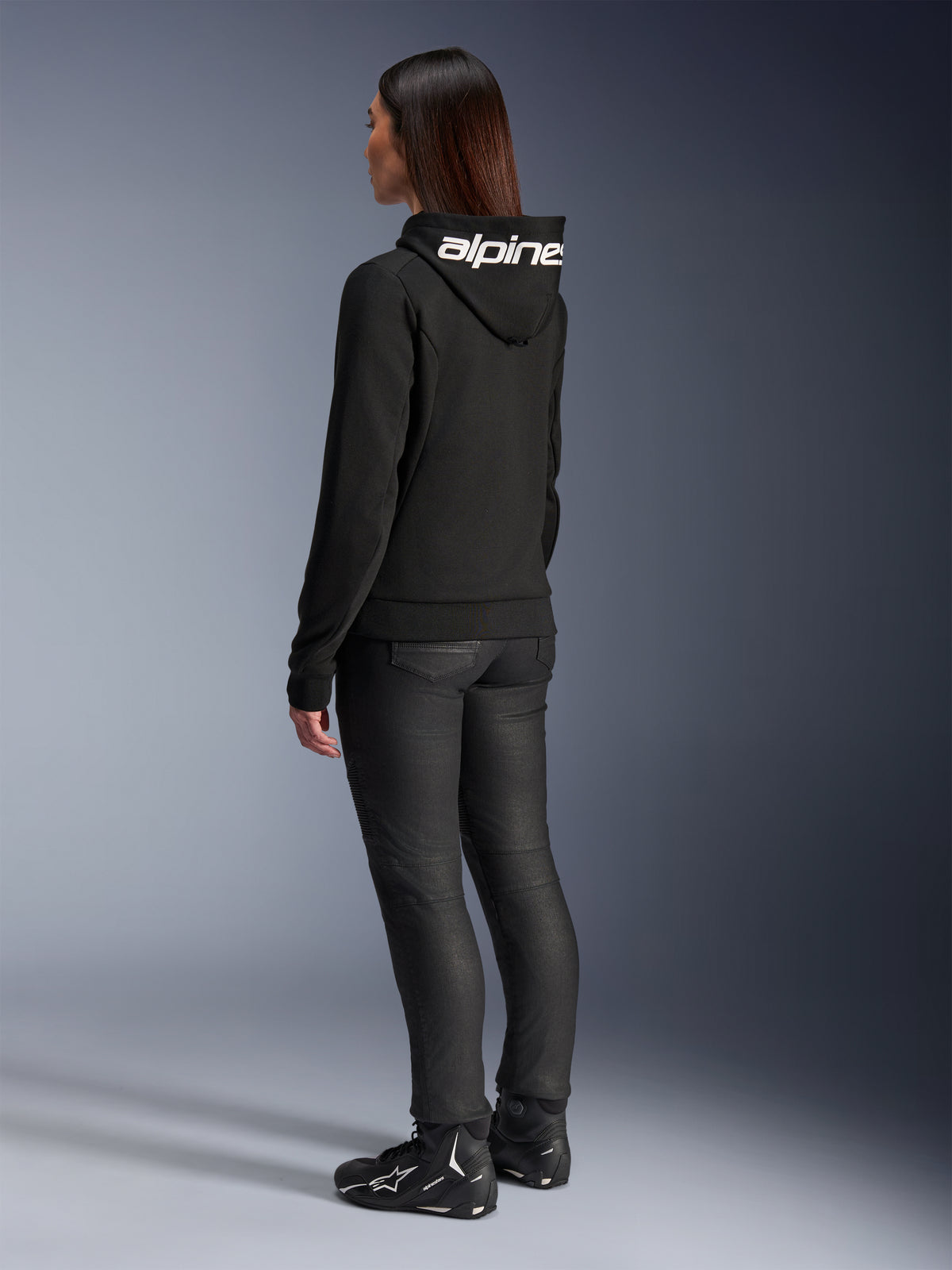 Donna che indossa la felpa Alpinestars Stella Chrome V2 Sport Hoodie, vista posteriore, felpa da moto nera con logo Alpinestars bianco sul cappuccio, abbinata a pantaloni tecnici da guida neri e scarpe da moto nere, abbigliamento casual per il pendolarismo urbano