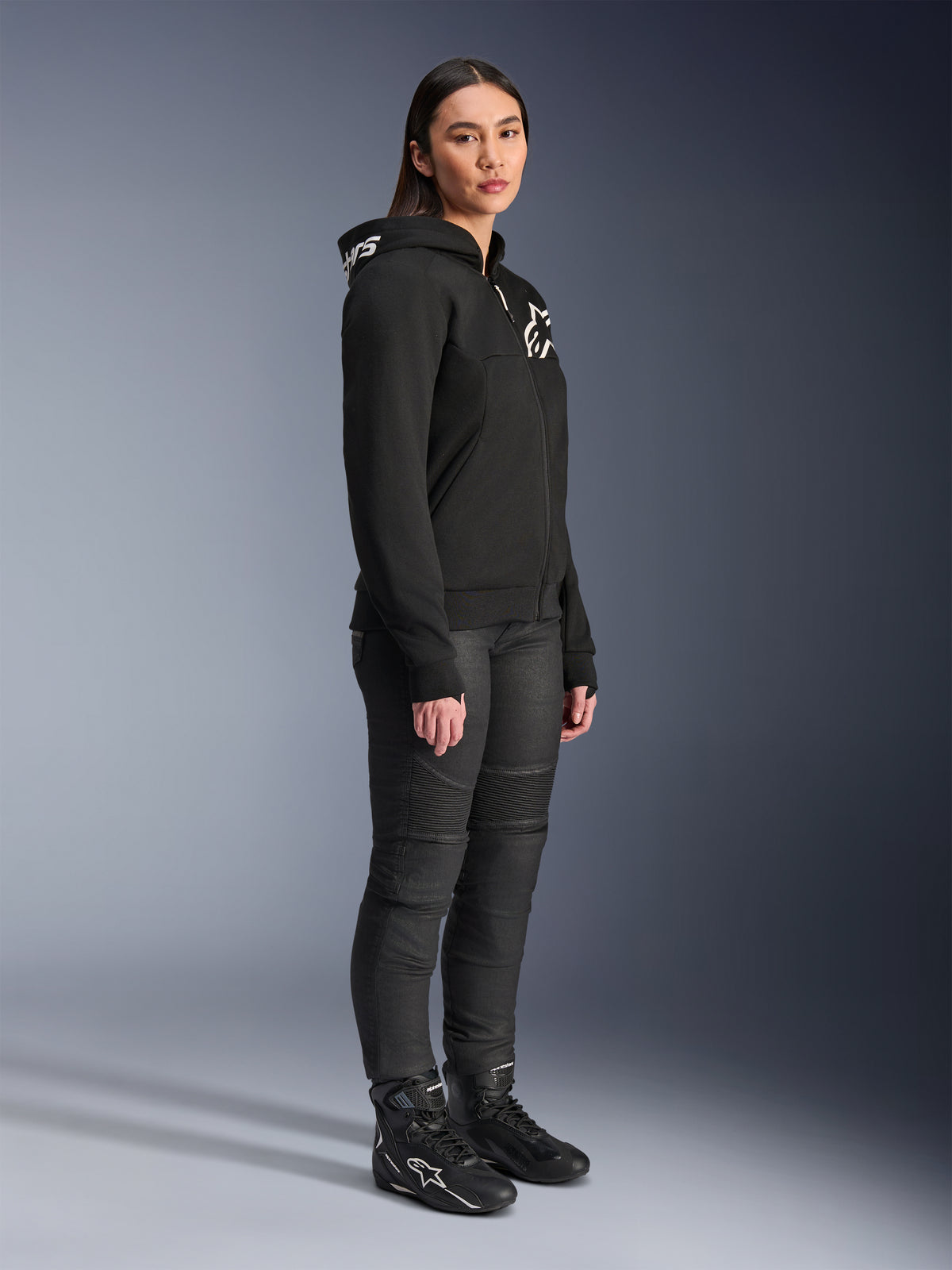 Donna che indossa la felpa Alpinestars Stella Chrome V2 Sport Hoodie in nero, con logo Alpinestars bianco, abbinata a pantaloni e stivali da moto neri, abbigliamento urban da donna con vestibilità atletica casual