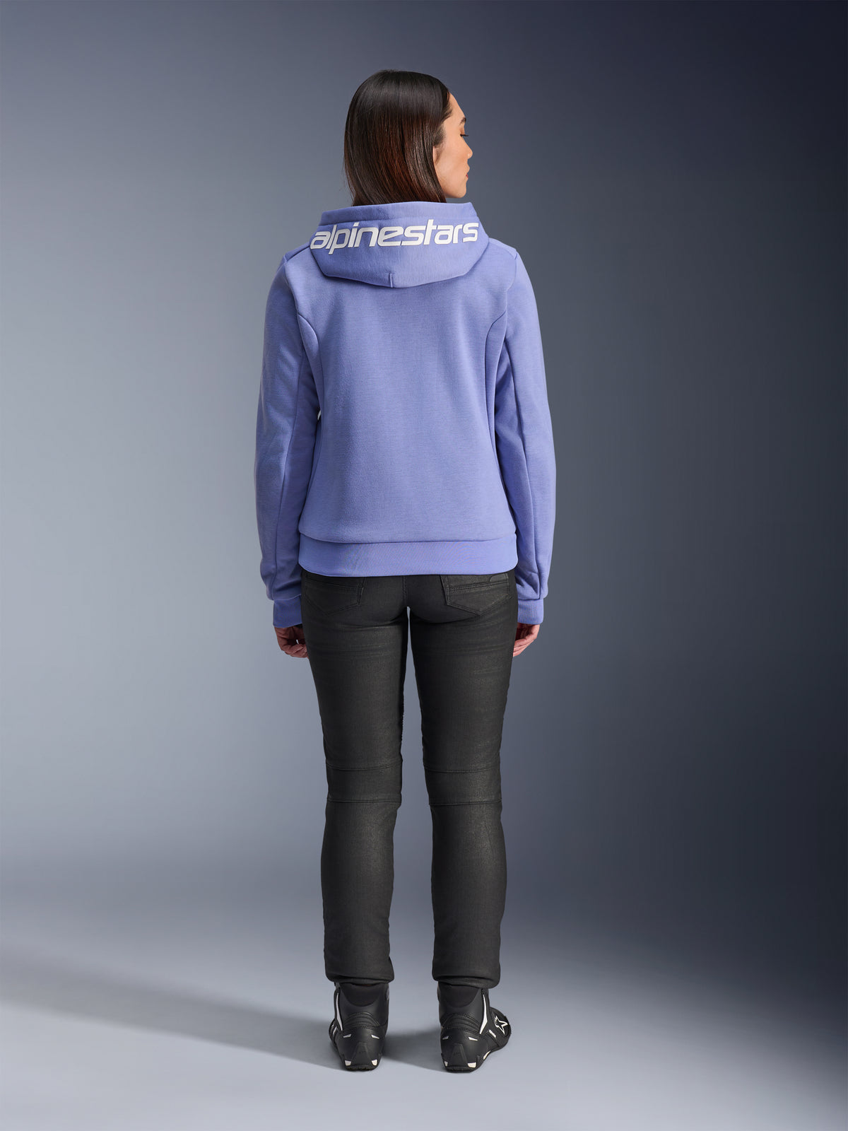 Alpinestars Stella Chrome V2 Sport Hoodie, blu violetto, vista posteriore di una donna che indossa la felpa con logo Alpinestars bianco sul cappuccio, abbinata a pantaloni neri e stivali da moto, abbigliamento casual da moto urbano