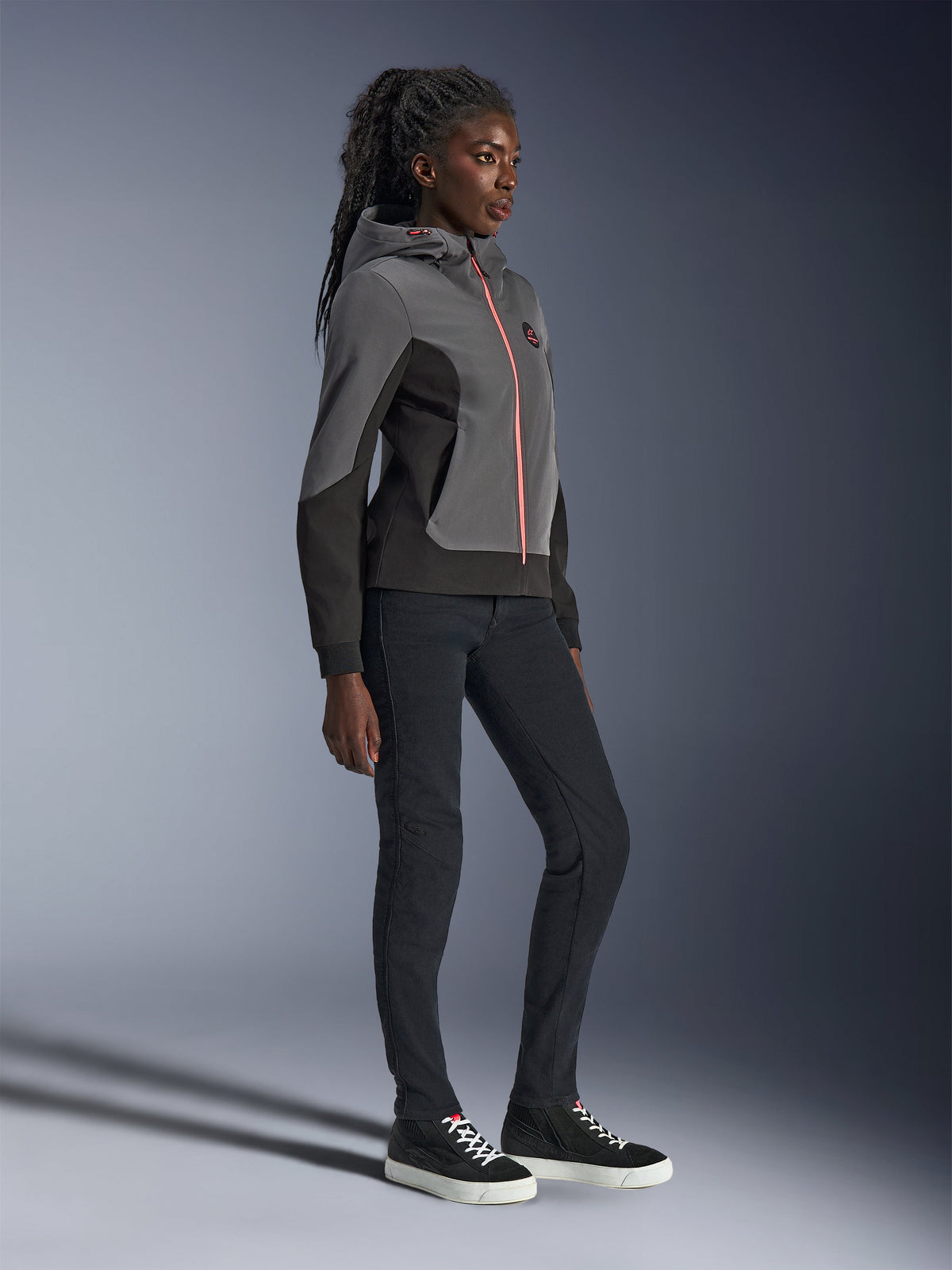 Alpinestars Stella Radium Tech Hoodie, felpa tecnica da moto urbana, grigio scuro e nero con cerniera arancione vivace, indossata con pantaloni neri e sneakers nere, vestibilità atletica specifica per donna