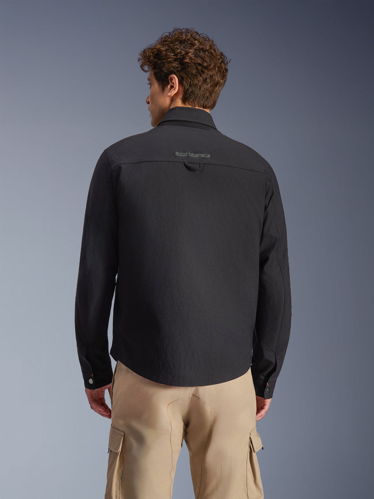 Alpinestars Bryon Water Resistant Overshirt, nero, vista posteriore indossata da un modello, con logo ricamato sullo sprone posteriore e asola di sospensione, abbinata a pantaloni cargo color sabbia, abbigliamento da moto urbano con estetica workwear classica