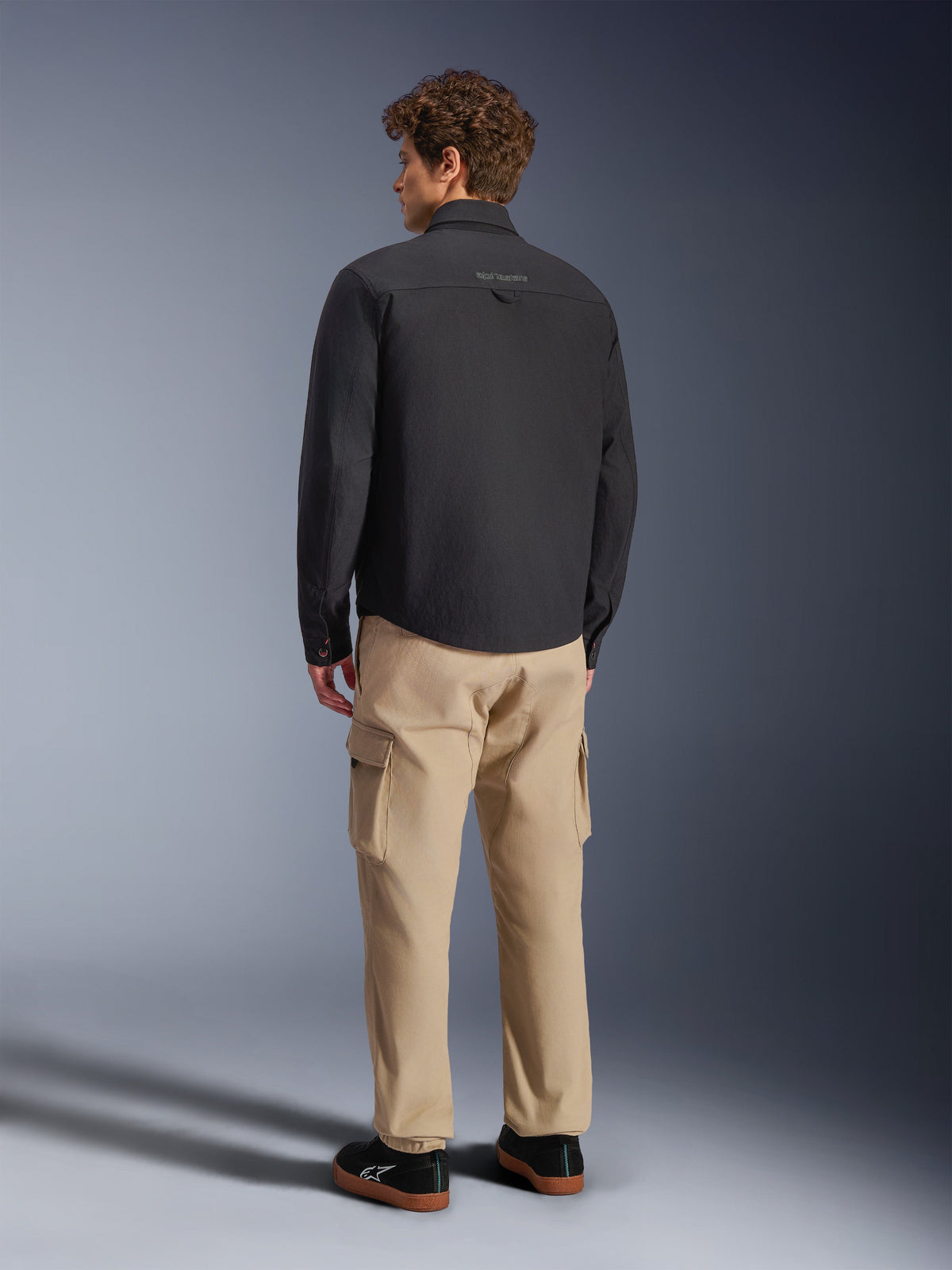 Alpinestars Bryon WR Overshirt, nero, vista posteriore su modello, abbinata a pantaloni cargo color sabbia e sneakers nere, giacca da moto urbana con texture in tela cerata e design posteriore minimalista