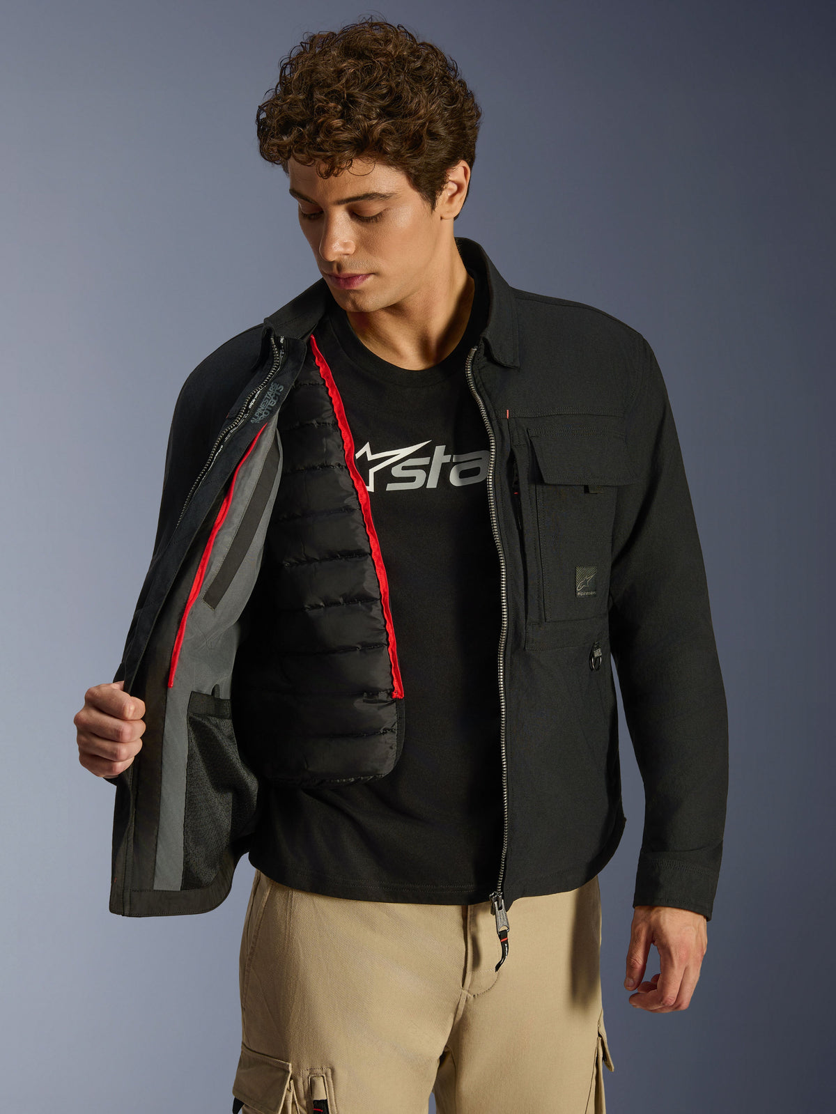 Alpinestars Bryon WR Overshirt, nero, giacca da moto urban mostrata aperta per rivelare un gilet termico trapuntato nero rimovibile con profili rossi, indossata con pantaloni cargo color sabbia e t-shirt con logo, design funzionale ispirato all'abbigliamento da lavoro