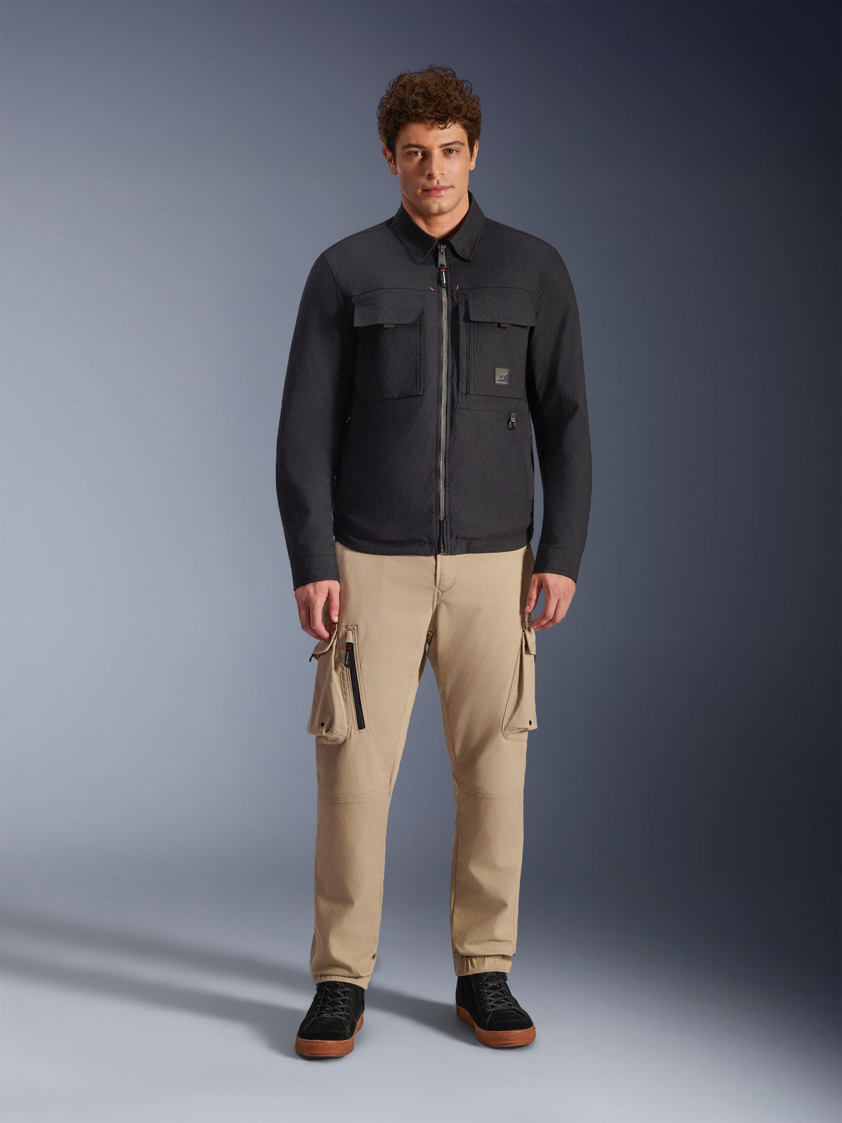 Modello che indossa l'overshirt Alpinestars Bryon Water Resistant in nero, con tasche cargo sul petto e cerniera centrale, abbinata a pantaloni cargo color sabbia e sneaker nere, ritratto in studio a figura intera.