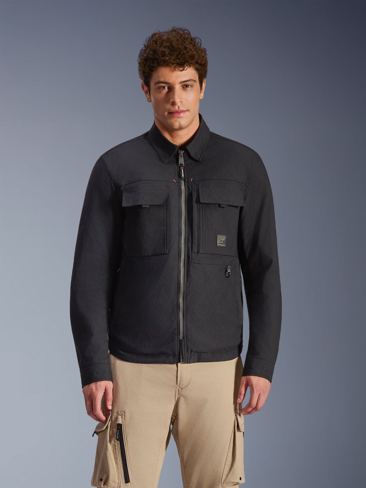 Alpinestars Bryon Water Resistant Overshirt, nero, giacca da moto urban dallo stile ispirato all'abbigliamento da lavoro, con due ampie tasche cargo sul petto e zip a tutta lunghezza, realizzata in canvas cerato, indossata da un modello con pantaloni cargo beige per un look utility contemporaneo.