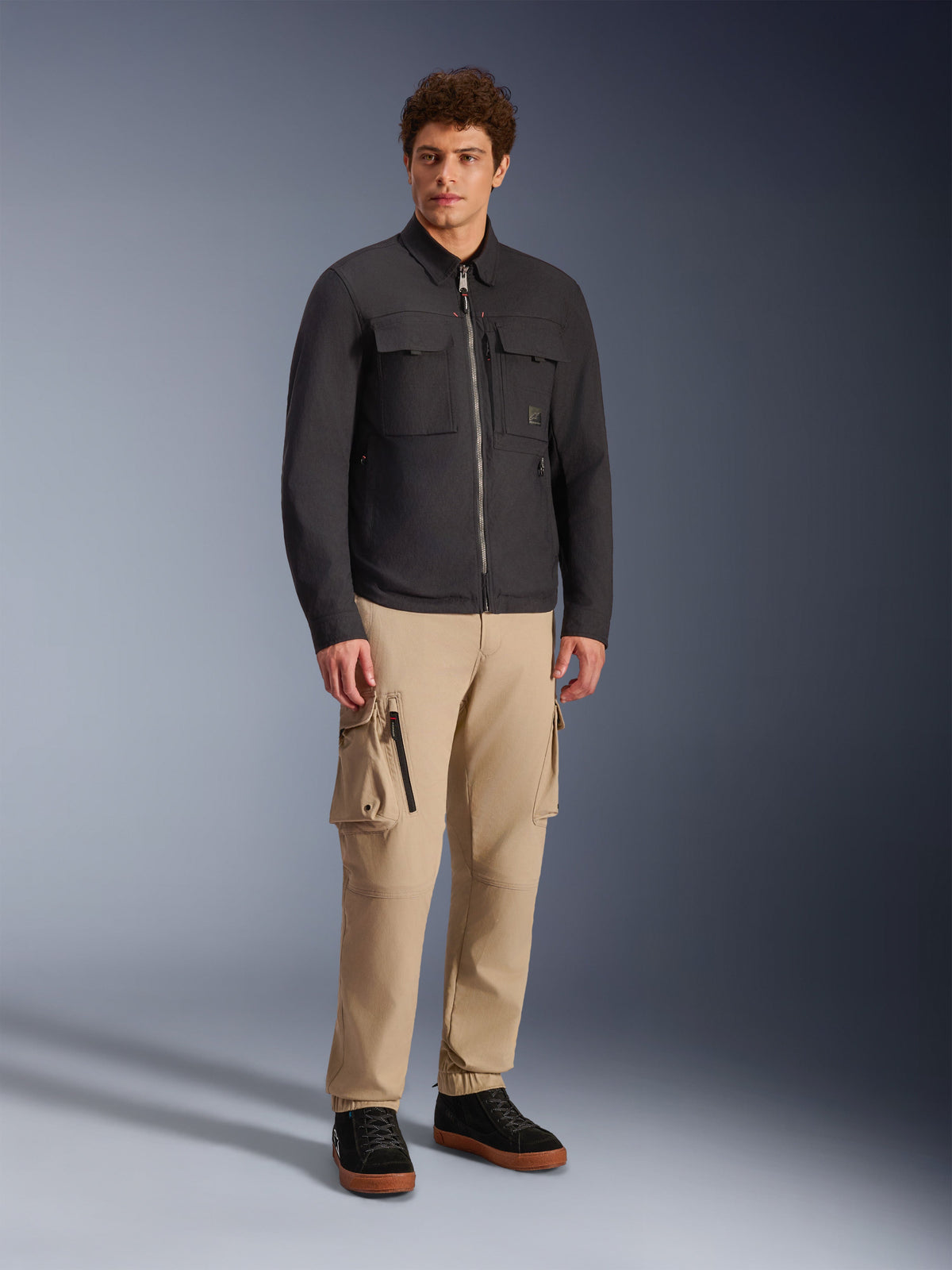 Alpinestars Bryon Water Resistant Overshirt, nero, indossata da un modello con pantaloni cargo kaki e scarpe nere, giacca da moto urbana dal design ispirato al workwear, scatto in studio a figura intera