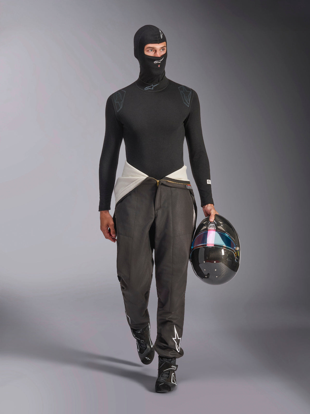 Passamontagna Alpinestars ZX Evo V3, nero, indossato da un modello con un completo base layer da corsa coordinato, maglia a maniche lunghe e pantaloni grigi, mentre tiene un casco in fibra di carbonio con visiera iridescente, equipaggiamento professionale per corse automobilistiche