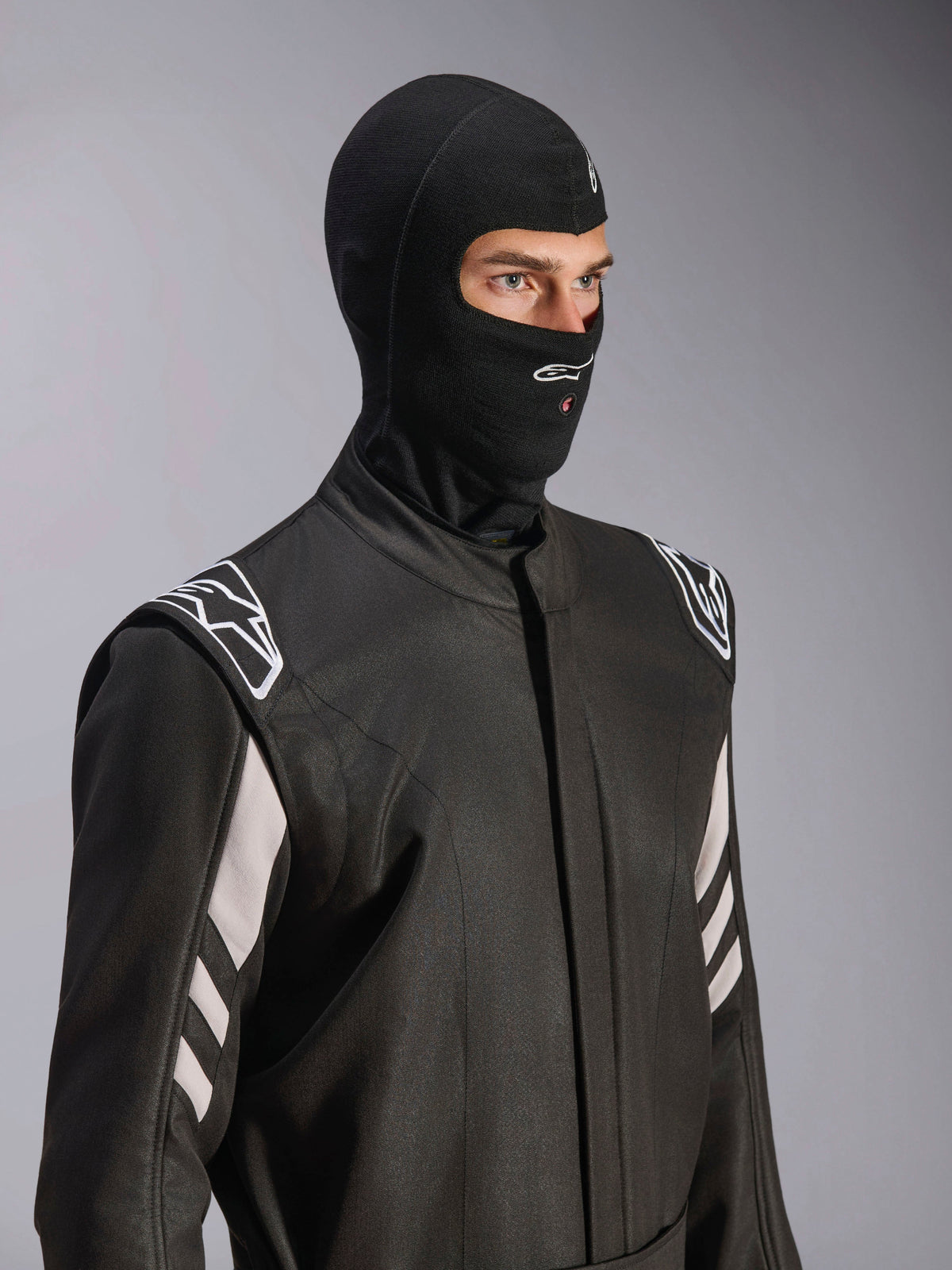 Alpinestars ZX Evo V3 Balaclava, nero, passamontagna ignifugo per corse automobilistiche, indossato da un modello in tuta da corsa nera con accenti bianchi, logo Alpinestars sul mento, apertura per il tubo di idratazione, equipaggiamento protettivo professionale per motorsport