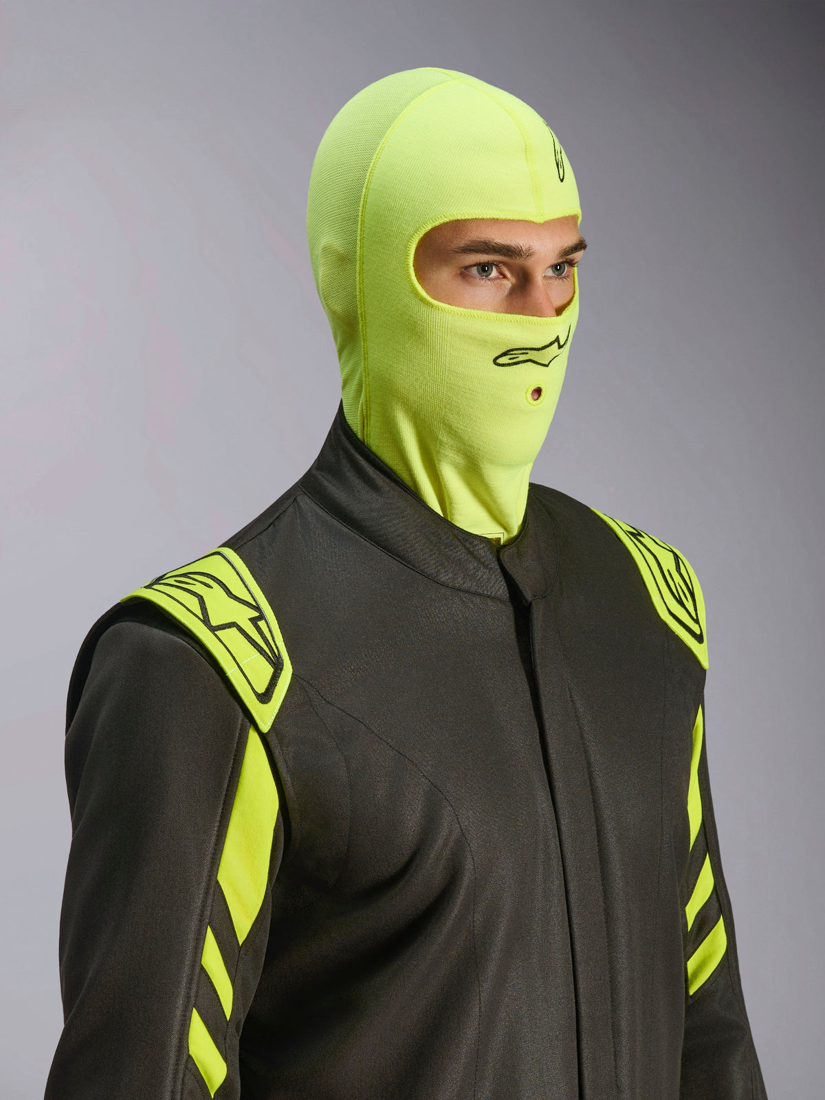 Modello che indossa il passamontagna Alpinestars ZX Evo V3 in Yellow Fluo, sottocasco da gara resistente alle fiamme, abbinato a una tuta da auto grigio scuro con accenti giallo neon, logo Alpinestars nero e apertura per tubo di idratazione, equipaggiamento protettivo professionale per motorsport.