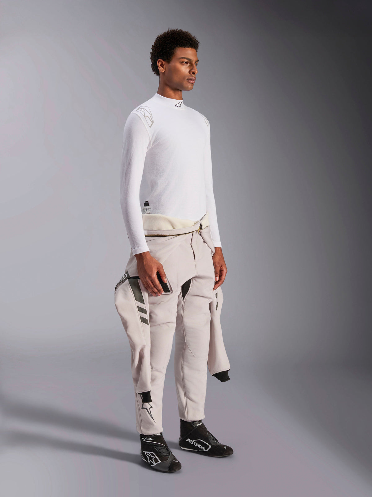 Alpinestars ZX Evo V3 Top a maniche lunghe, base layer per auto racing bianco resistente alle fiamme, indossato da un modello con una tuta da gara legata in vita, strato tecnico leggero con collo a lupetto e dettaglio del logo.