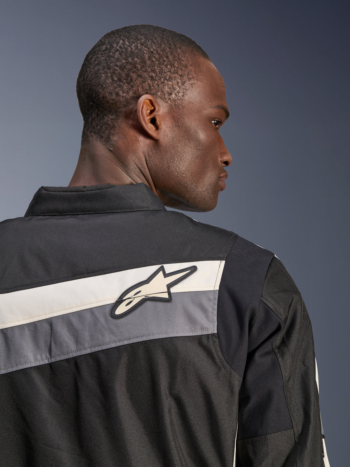 Giacca Alpinestars T-Dyno WR, giacca da moto in tessuto, nero, grigio scuro ed ecru, primo piano della parte superiore della schiena e della spalla con strisce orizzontali e logo a stella Alpinestars, indossata da un modello, design di ispirazione heritage per la guida su strada