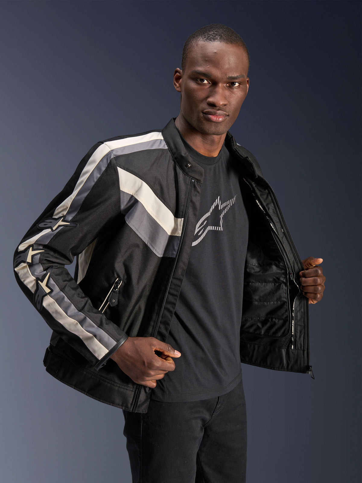 Giacca Alpinestars T-Dyno WR, Nero Grigio Scuro ed Ecru, giacca da moto in tessuto con vestibilità da biker d'ispirazione vintage, dettagli a strisce su petto e maniche, indossata sopra una t-shirt grafica nera, abbigliamento urban riding