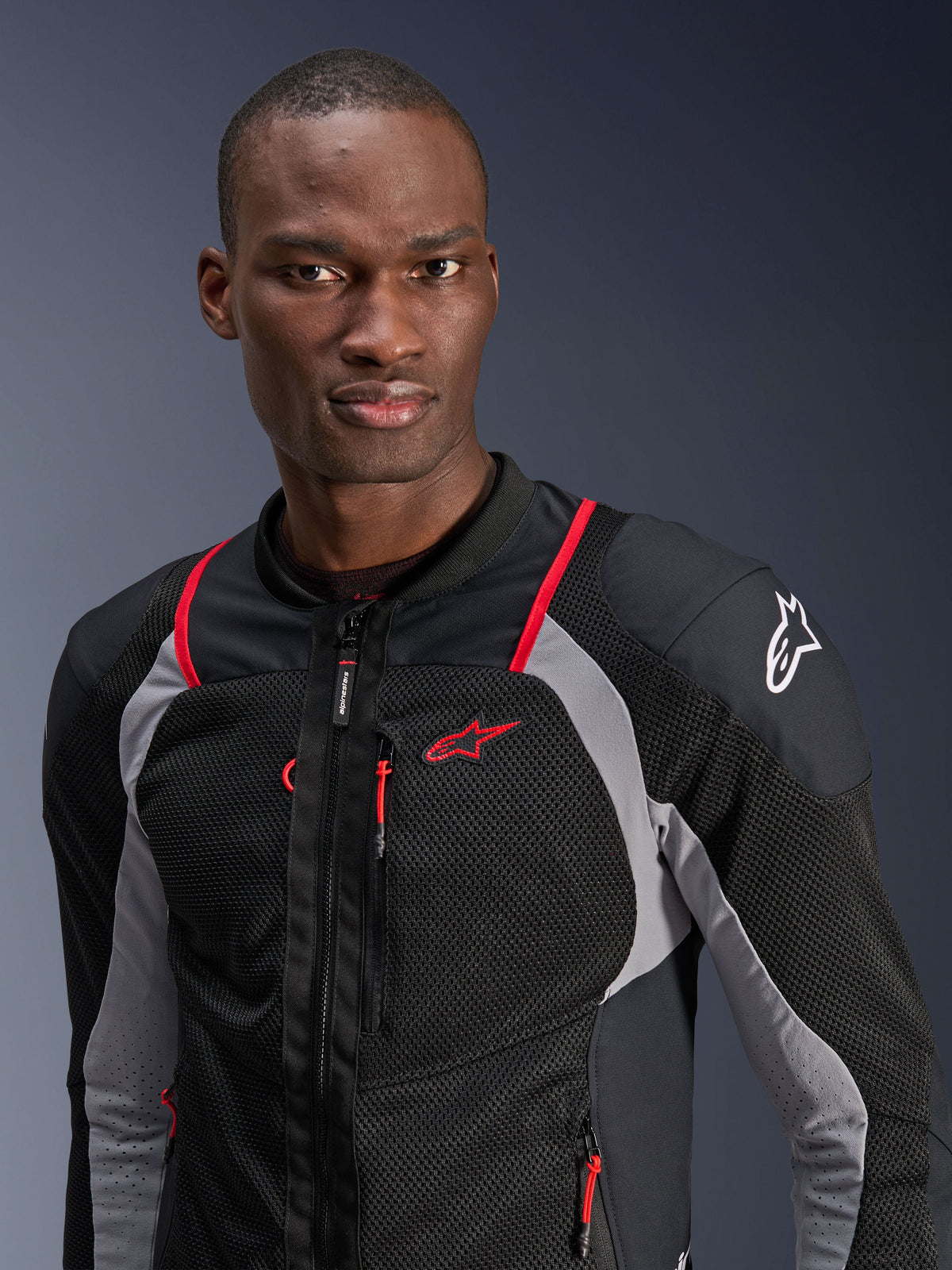 Uomo che indossa la giacca Alpinestars Troop-Air Pro, nero, grigio cenere e rosso brillante, giacca tecnica corazzata con pannelli in mesh traspirante, profili rossi e logo Alpinestars sulla spalla, abbigliamento da moto urban contemporary