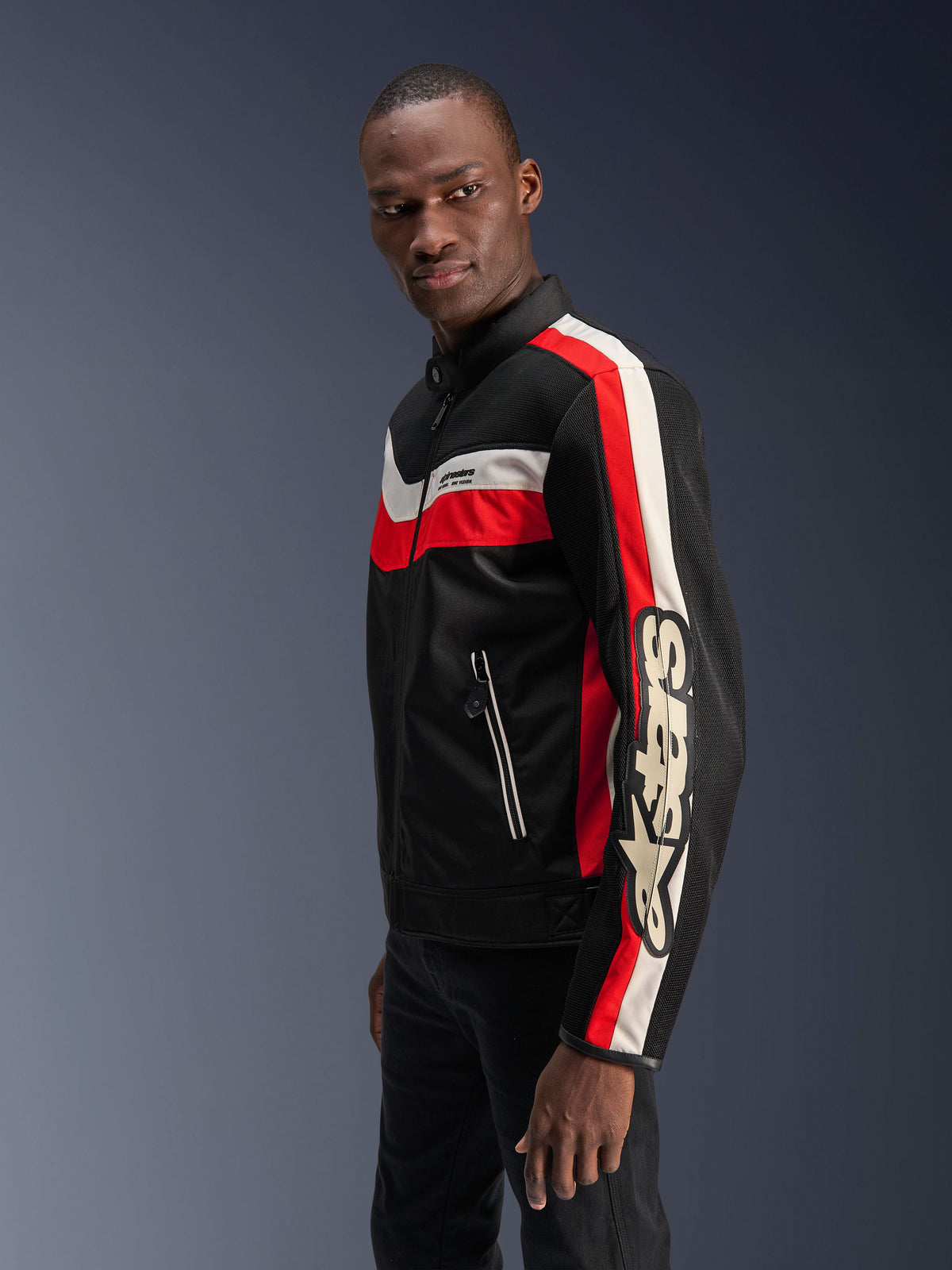 Modello uomo che indossa la giacca Alpinestars T-Dyno Air in nero, rosso acceso ed ecru, giacca da moto in tessuto d'ispirazione vintage con pannelli in mesh 3D, audaci strisce rosse e bianco sporco e marchio Alpinestars sulla manica.