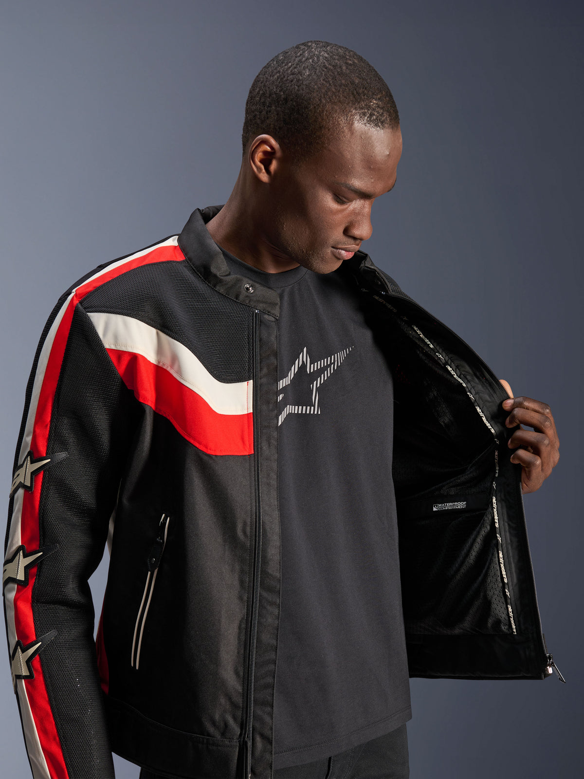 Giacca Alpinestars T-Dyno Air, Nero Rosso Brillante ed Ecru, modello maschile che apre la giacca per mostrare la fodera interna e lo scomparto impermeabile, giacca in tessuto d'ispirazione vintage con pannelli in rete 3D e loghi a stella, moderno abbigliamento tecnico da moto