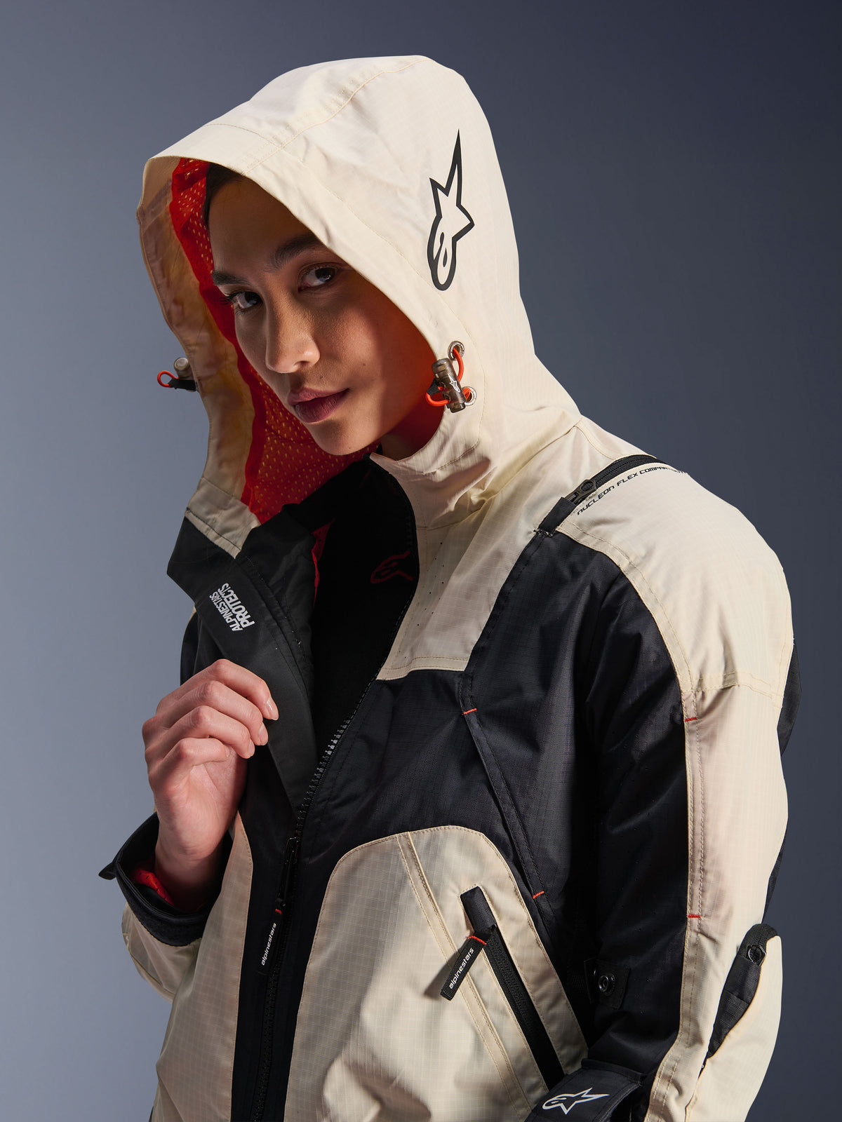 Giacca Alpinestars Molly Air in Black Almond, indossata da una modella con il cappuccio sollevato, caratterizzata da un design color-block crema e nero, fodera in rete arancione vivace ed elementi di design tecnico urbano con logo a stella e dettagli con cerniera.