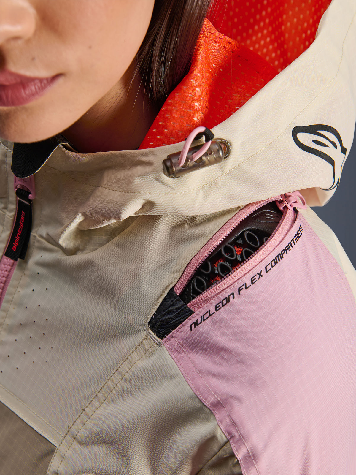 Primo piano della giacca Alpinestars Molly Air in Sand Pink Almond, che mette in risalto lo scomparto sulla spalla con zip per le protezioni Nucleon Flex, la fodera del cappuccio in rete arancione brillante e la costruzione in resistente tessuto ripstop.