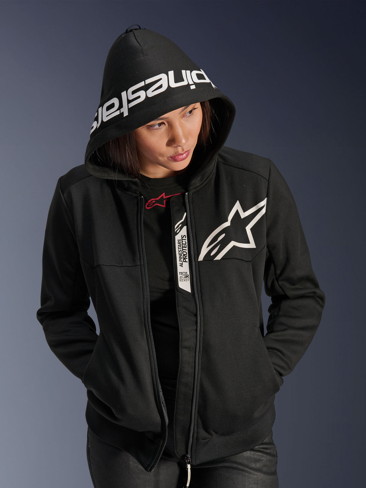 Alpinestars Stella Chrome V2 Sport Hoodie da donna, nero e bianco, felpa da moto con cappuccio, logo a stella bianco sul petto e scritta Alpinestars sul bordo del cappuccio, modella con cappuccio alzato, stile sportivo urbano casual con dettagli Tech-Air Ready