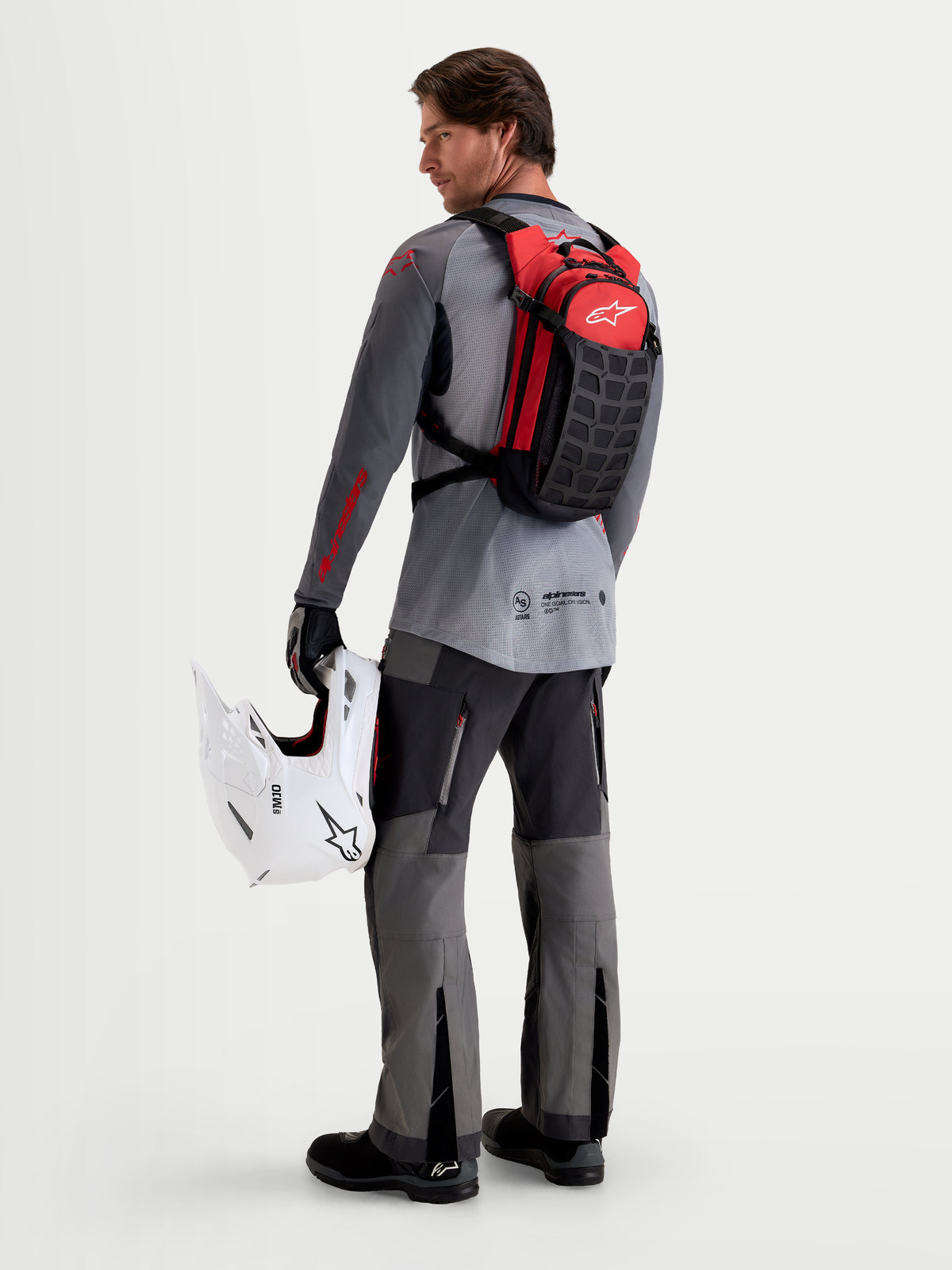 Alpinestars Zaino AT-9, zaino da motocross nero e rosso acceso, indossato da un modello di spalle che tiene un casco MX bianco e attrezzatura coordinata, struttura leggera in tessuto per l'uso in pista e nel paddock