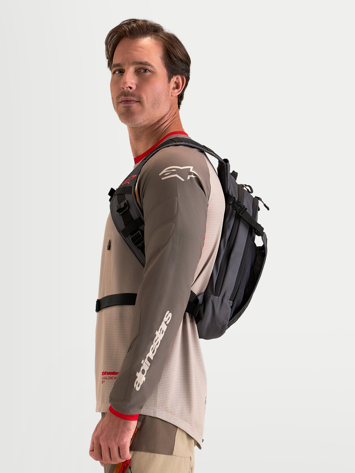 Alpinestars Zaino AT-9, zaino da motocross in marrone noce e grigio scuro, indossato da un modello che mostra il profilo dell'imbracatura laterale e la struttura in tessuto leggero