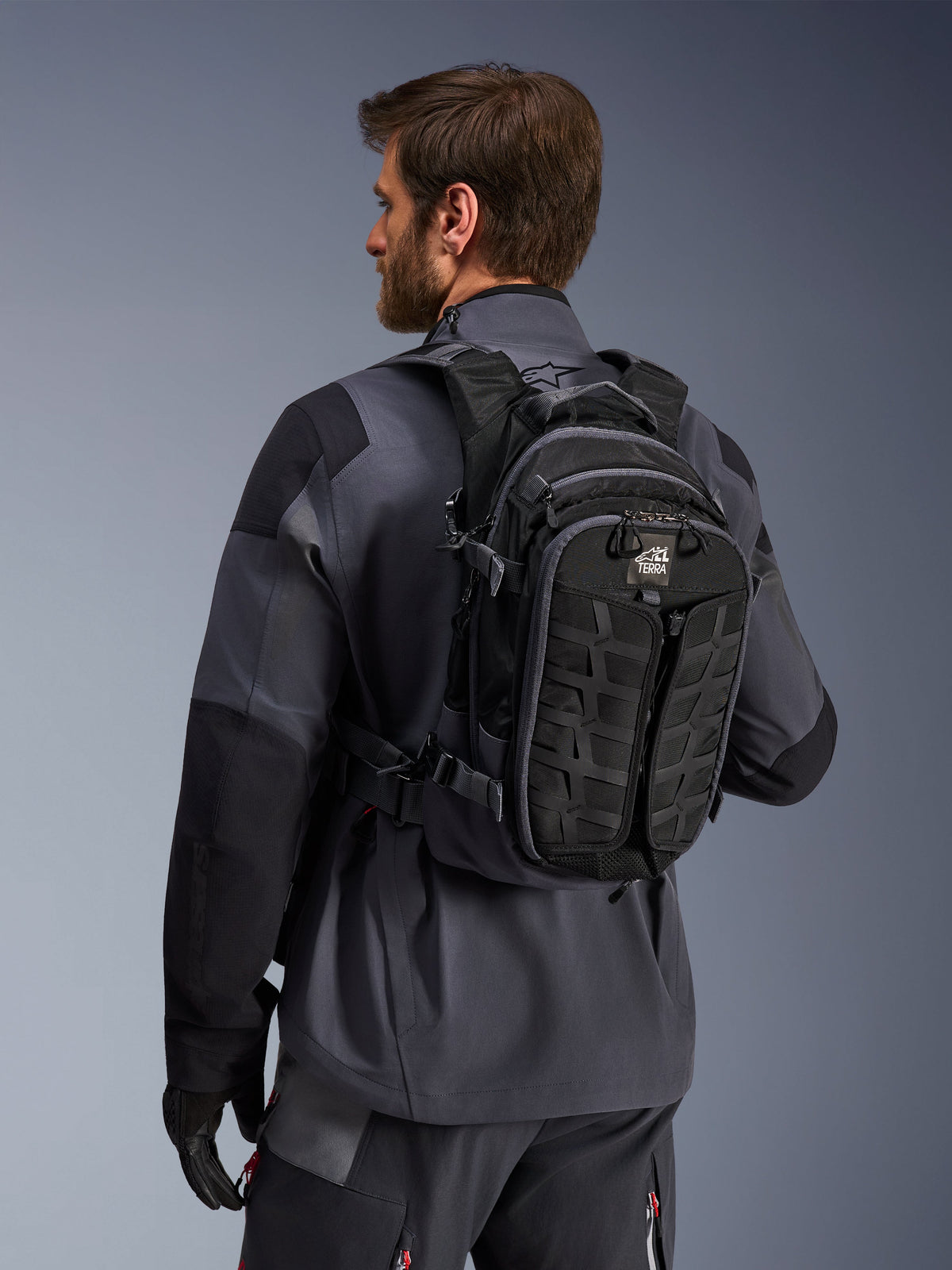 Alpinestars AT-15 Back Pack, zaino da moto adventure, nero e grigio scuro, indossato da un pilota con giacca grigia, caratterizzato da un design tecnico con sistema MOLLE e marchio 'TERRA', equipaggiamento ergonomico per il fuoristrada