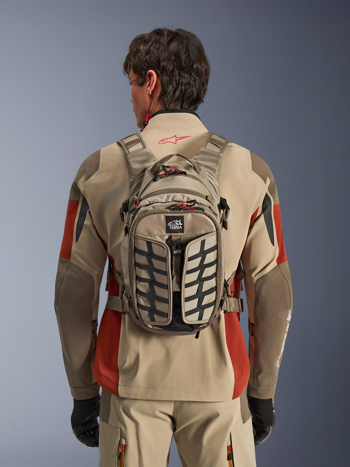 Alpinestars AT-15 Back Pack, colore Aluminum Walnut, zaino tecnico da moto adventure indossato con giacca beige e rossa, con motivo a impronta di pneumatico, logo TERRA, tiretti delle cerniere rossi e cinghie regolabili