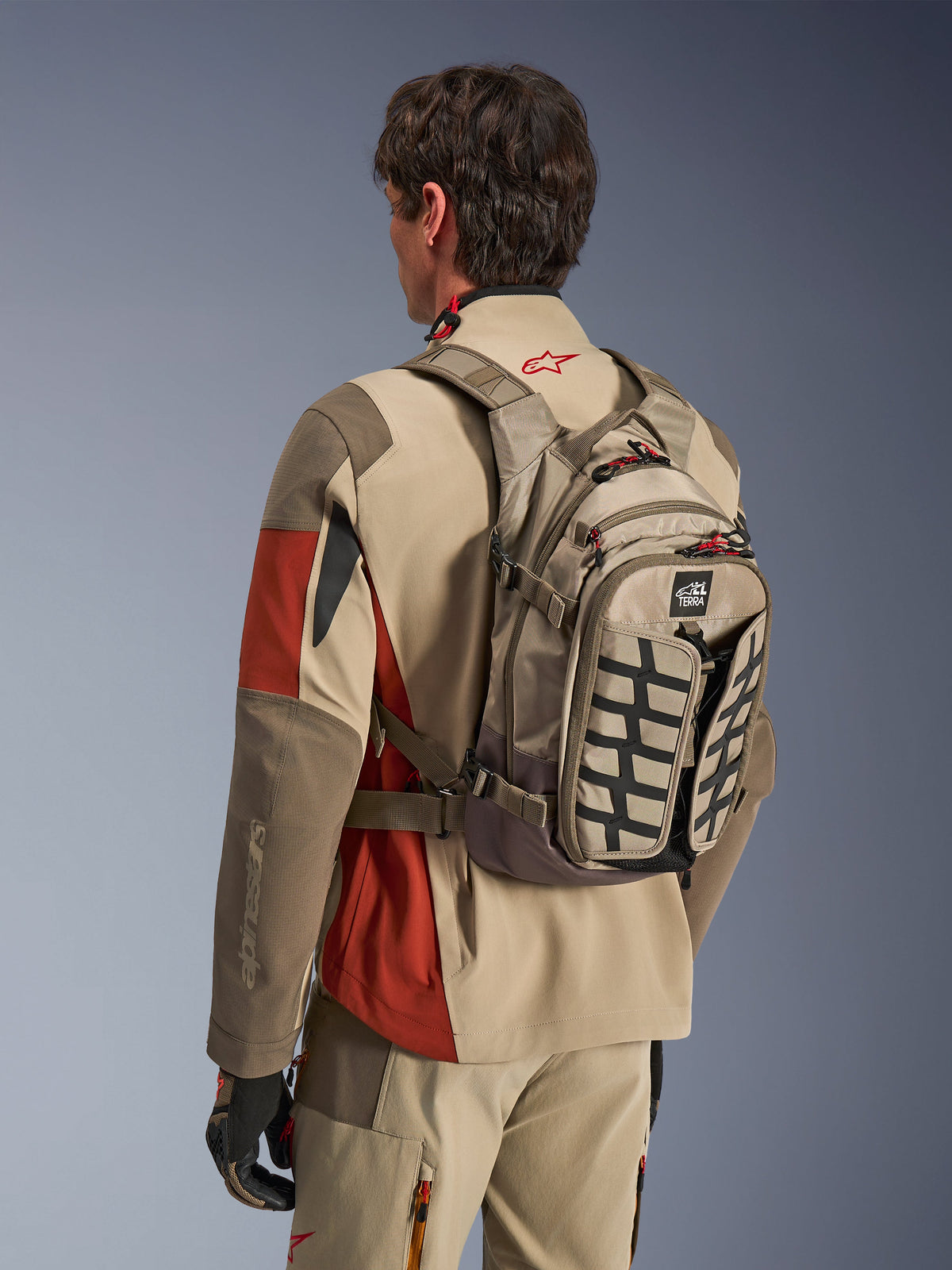 Zaino Alpinestars AT-15 Back Pack in Aluminum Walnut, zaino tecnico da moto indossato da un pilota con abbigliamento adventure coordinato, caratterizzato da una costruzione in tessuto tan e marrone con accenti strutturali neri e tiranti delle cerniere rossi, progettato per il fuoristrada e l'adventure riding.
