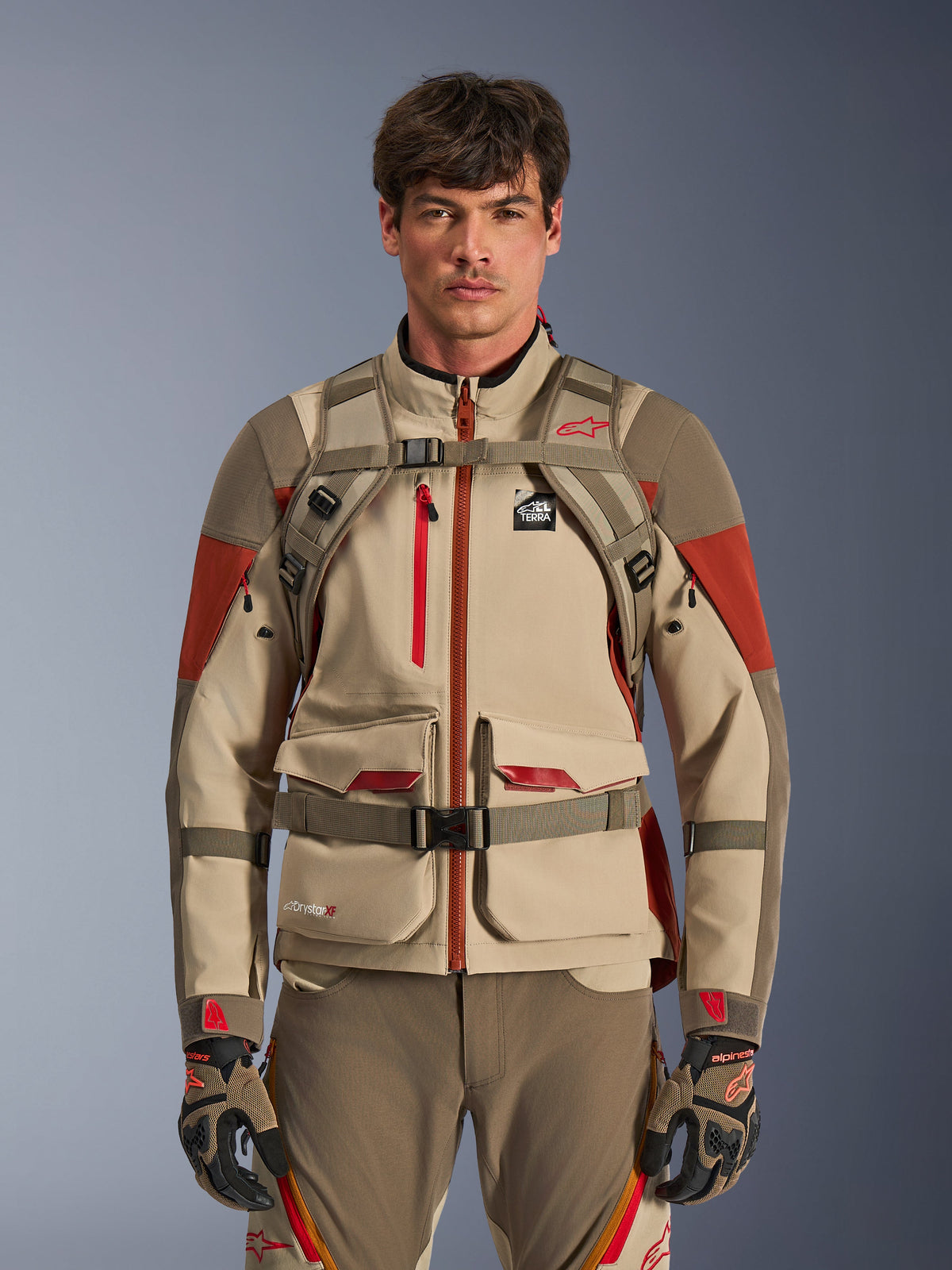 Pilota adventure che indossa lo zaino Alpinestars AT-15 in colore Aluminum Walnut, vista frontale con cinghie pettorali e addominali con chiusura magnetica, abbinato a giacca e pantaloni da moto adventure sabbia e rosso, completo tecnico da fuoristrada