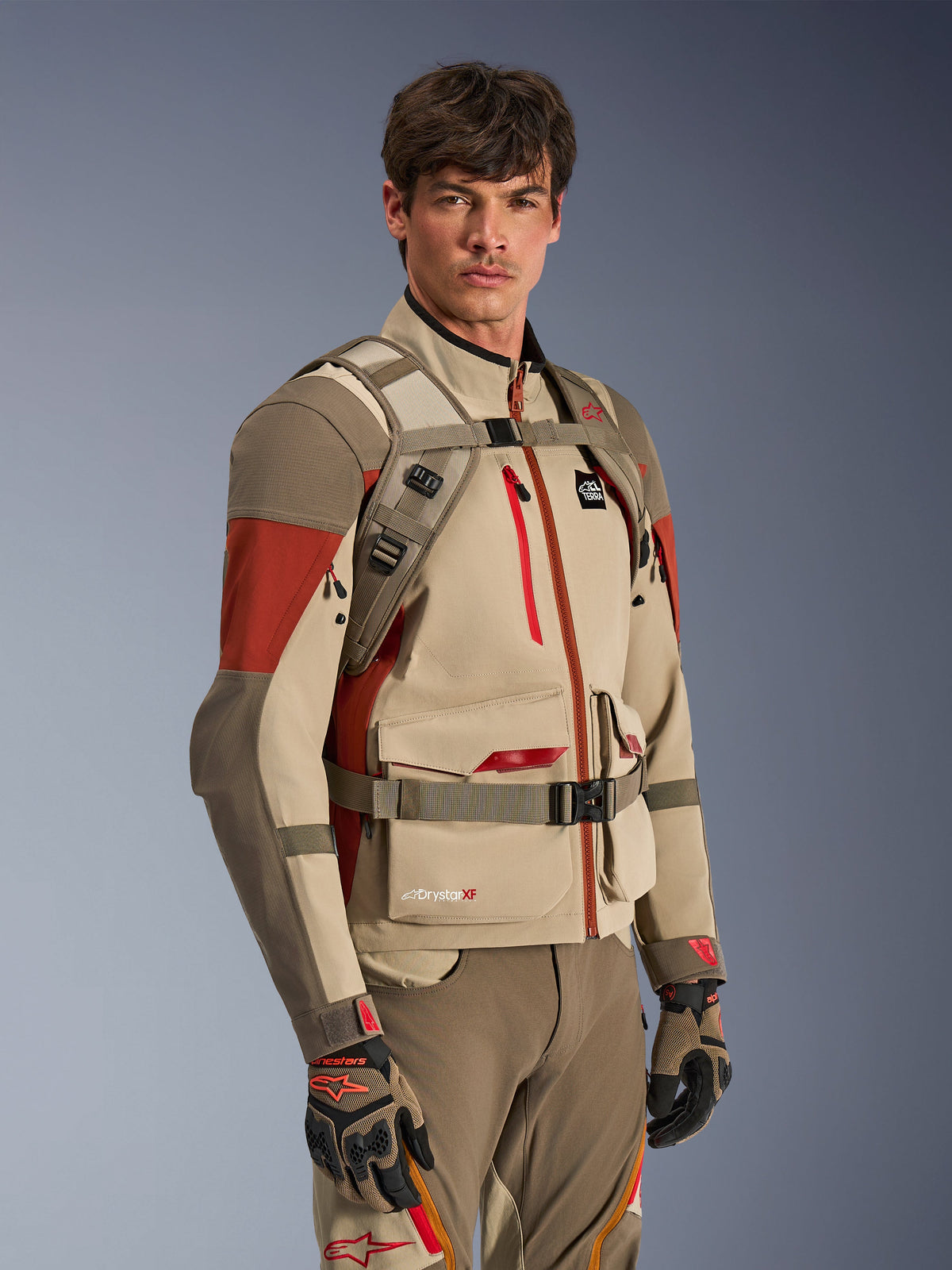 Zaino Alpinestars AT-15 in Aluminum Walnut, indossato da un pilota sopra una giacca da moto adventure abbinata, mostra l'imbracatura anteriore con chiusura magnetica sul petto e cinghie regolabili, abbigliamento tecnico beige e rosso per guida adventure