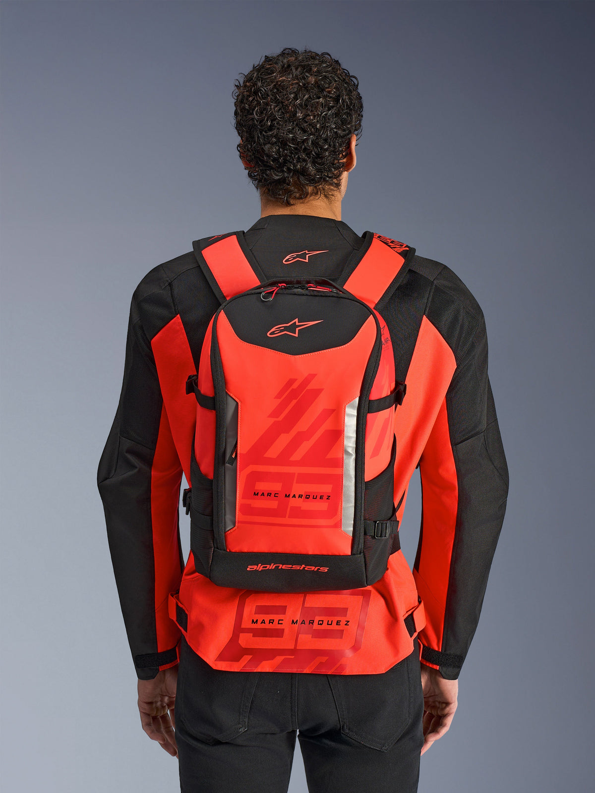 Alpinestars MM93 AMP3 Backpack, zaino tecnico da moto, rosso fluo e nero, indossato da un pilota visto da dietro sopra una giacca da corsa MM93 coordinata in rosso e nero, con marchio Marc Marquez e loghi Alpinestars.