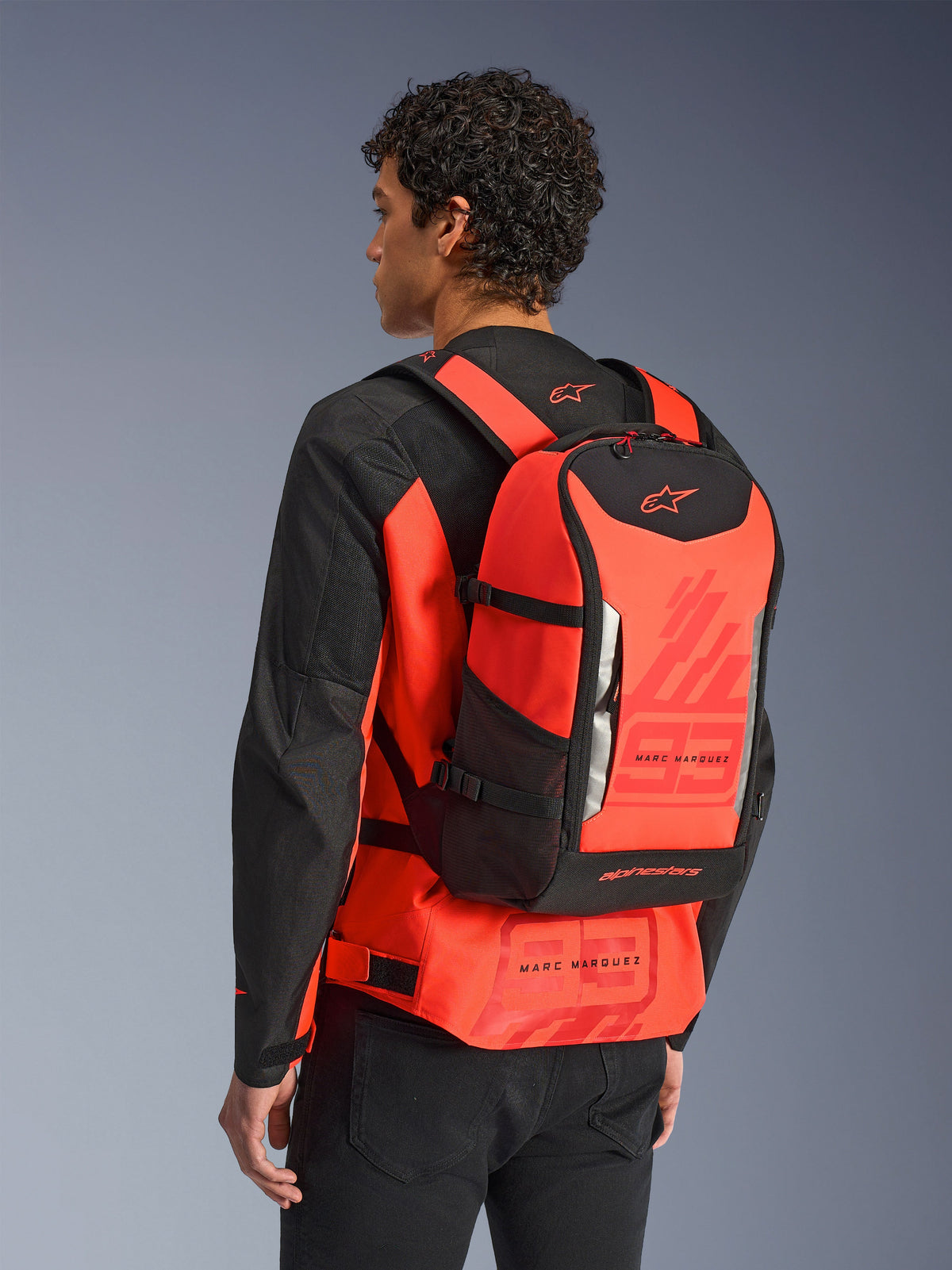 Zaino Alpinestars MM93 AMP3, zaino tecnico da moto, rosso fluo e nero, indossato da un modello visto da dietro sopra una giacca coordinata rossa e nera, con grafiche '93' di Marc Marquez, spallacci regolabili e design elegante di ispirazione sportiva.