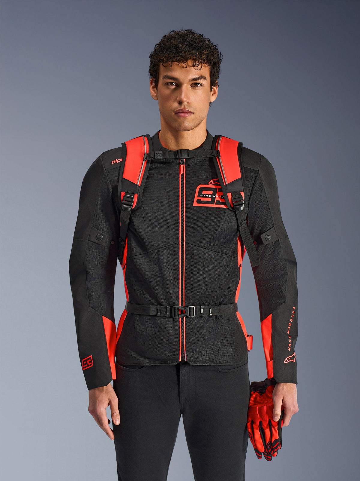 Uomo che indossa lo zaino Alpinestars MM93 AMP3, rosso fluo e nero, con spallacci regolabili e cinturino pettorale con fibbia, abbinato a giacca da moto e guanti MM93 coordinati, equipaggiamento tecnico della collezione Marc Marquez