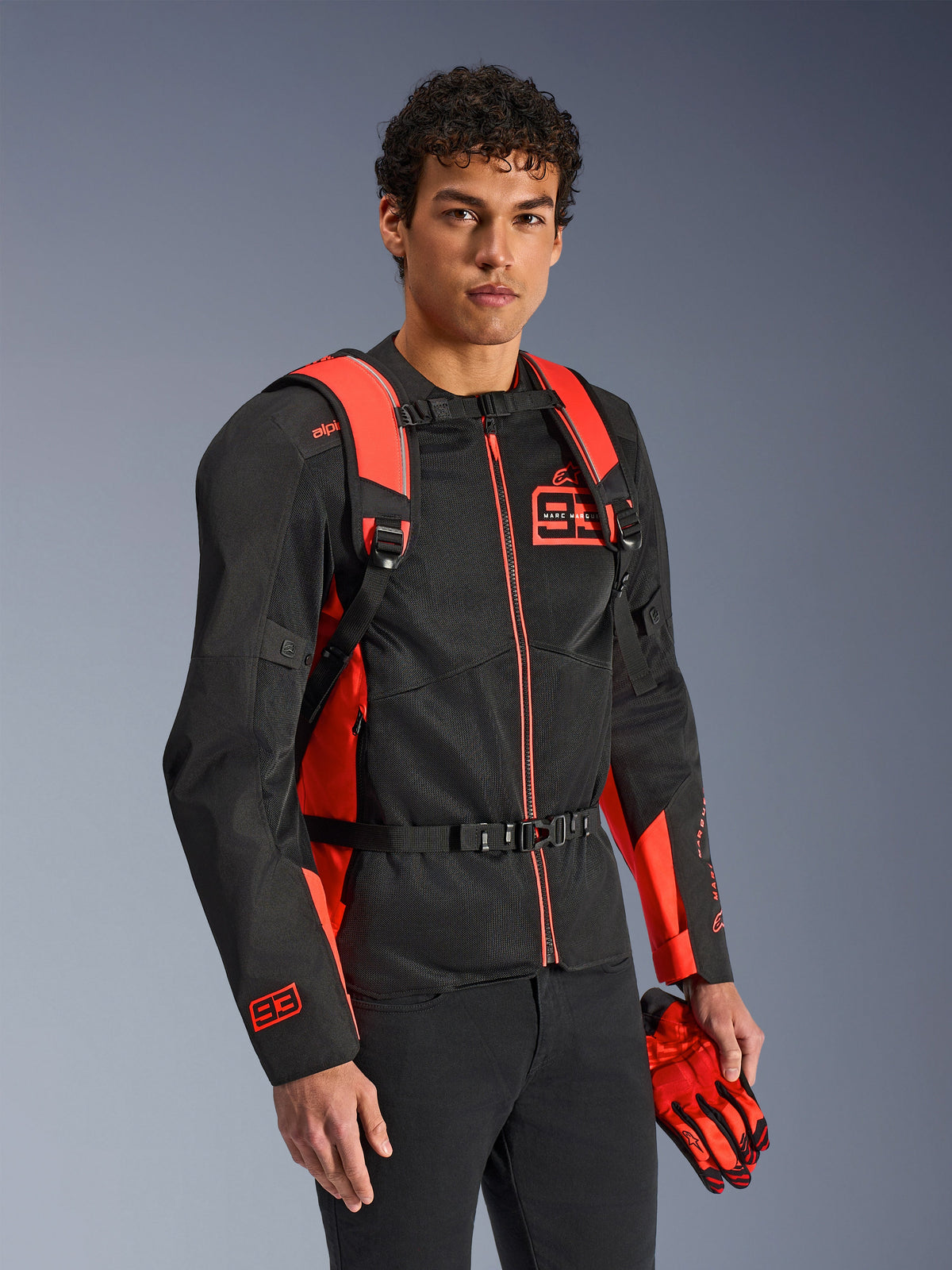 Zaino Alpinestars MM93 AMP3, rosso fluo e nero, indossato da un modello con giacca da moto e guanti MM93 coordinati neri e rossi, vista frontale che mette in risalto gli spallacci regolabili e la fibbia sul petto, equipaggiamento tecnico ispirato alle corse