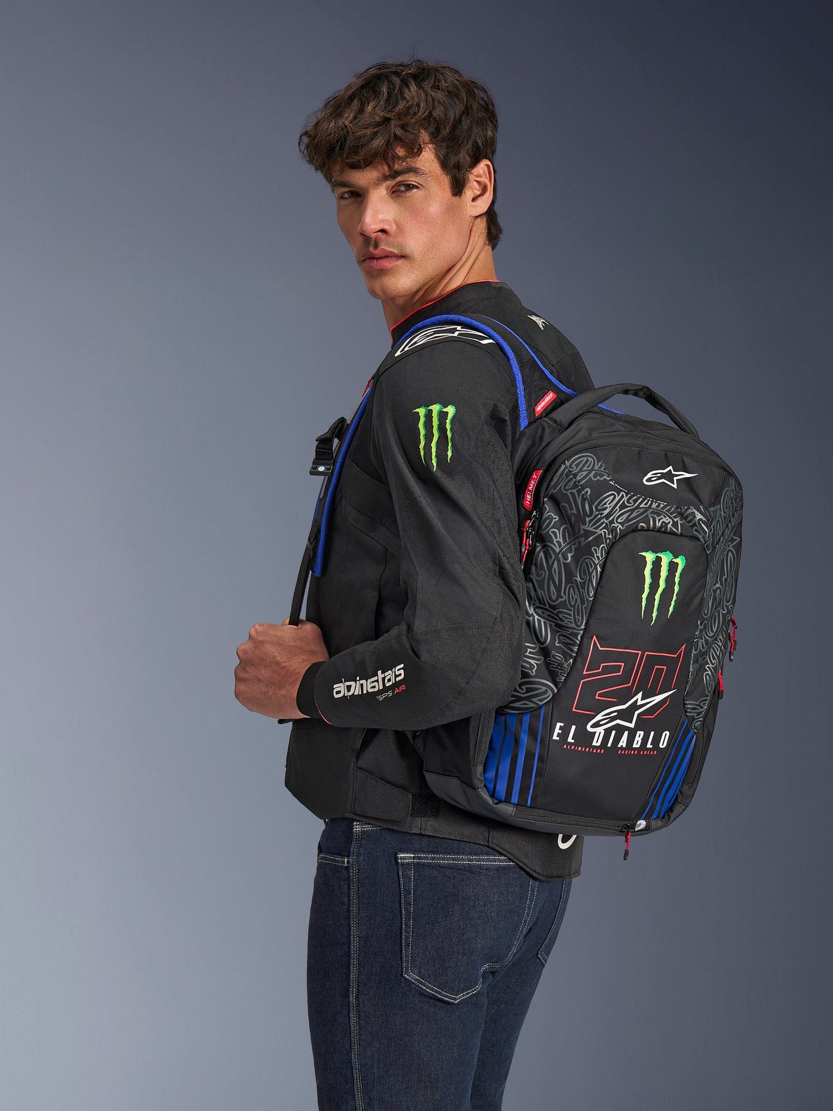 Alpinestars FQ20 City Hunter V2 Monster Backpack, zaino da moto, nero con rosso fluo, blu e verde, con branding Monster Energy e Fabio Quartararo 'El Diablo', indossato da un modello con giacca da moto nera e jeans, design aerodinamico per motociclisti urbani