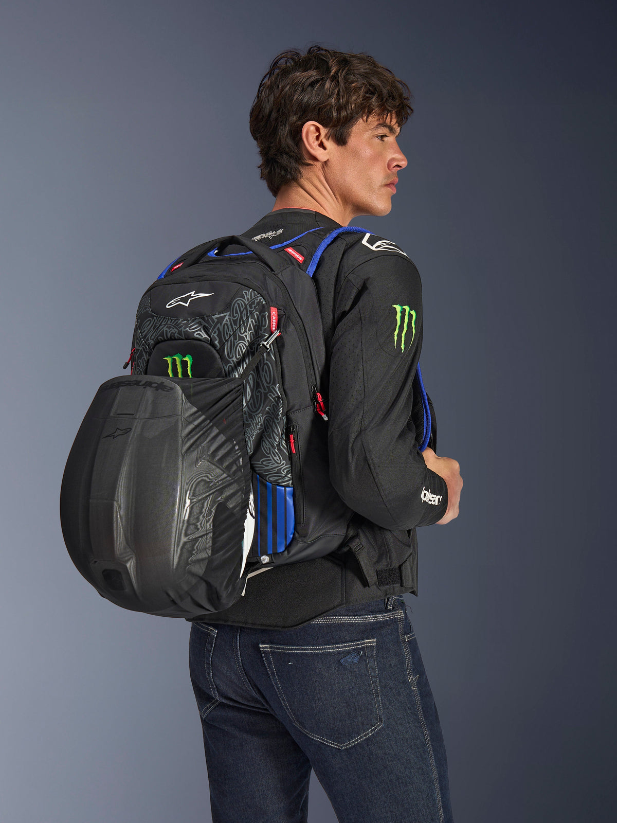 Alpinestars FQ20 City Hunter V2 Monster Backpack, zaino da moto urbano, nero con accenti rosso fluo, blu e verde, indossato da un modello con casco nero fissato nel porta-casco in rete posteriore, abbinato a giacca da moto nera e jeans blu