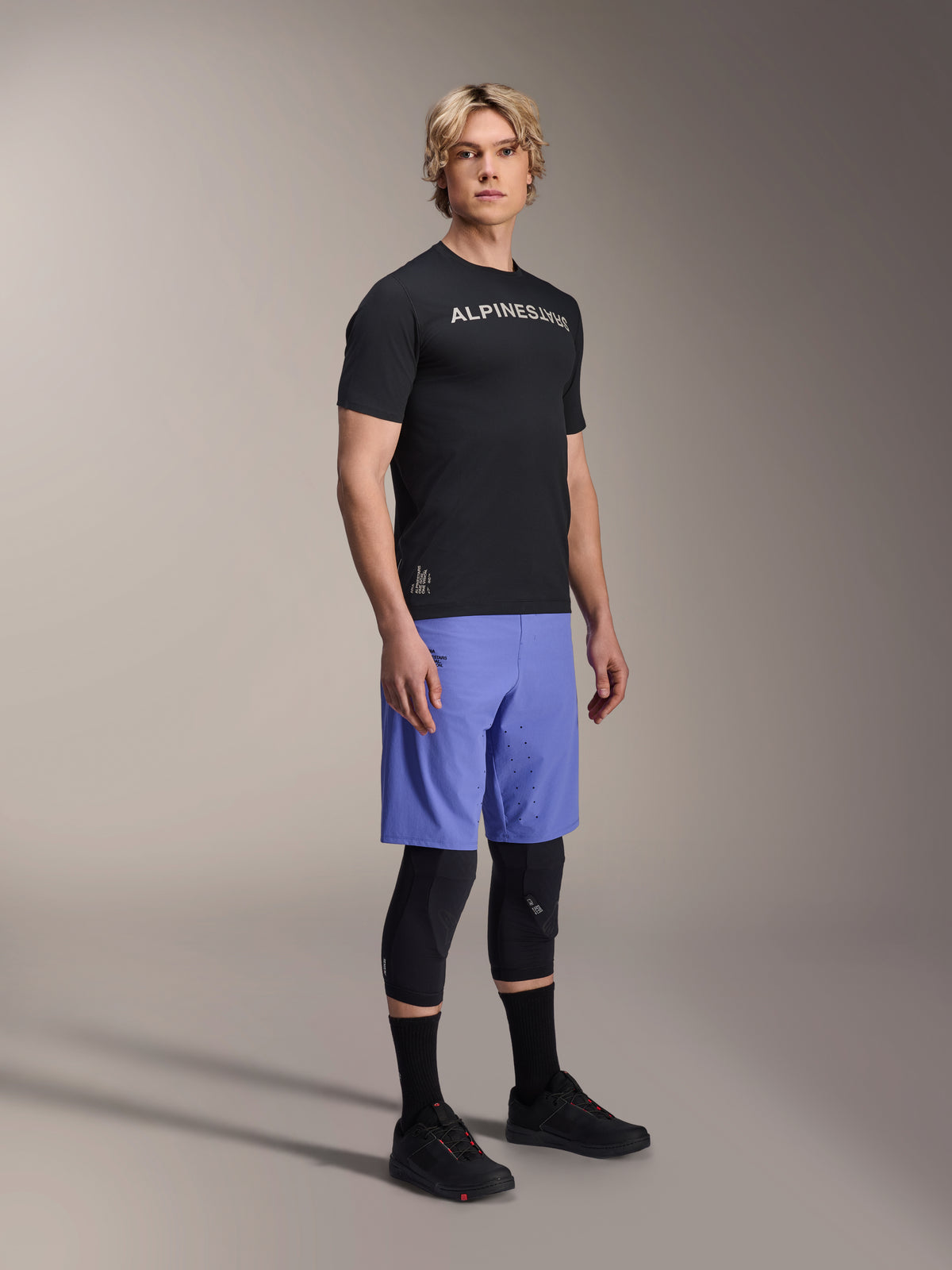 Modello che indossa abbigliamento da mountain bike Alpinestars, con la maglia a maniche corte A-Aria Switch nera e i pantaloncini A-Aria Lite Baja Blue, abbinati a ginocchiere nere, calze nere e scarpe da mountain bike nere in uno scatto in studio a figura intera.