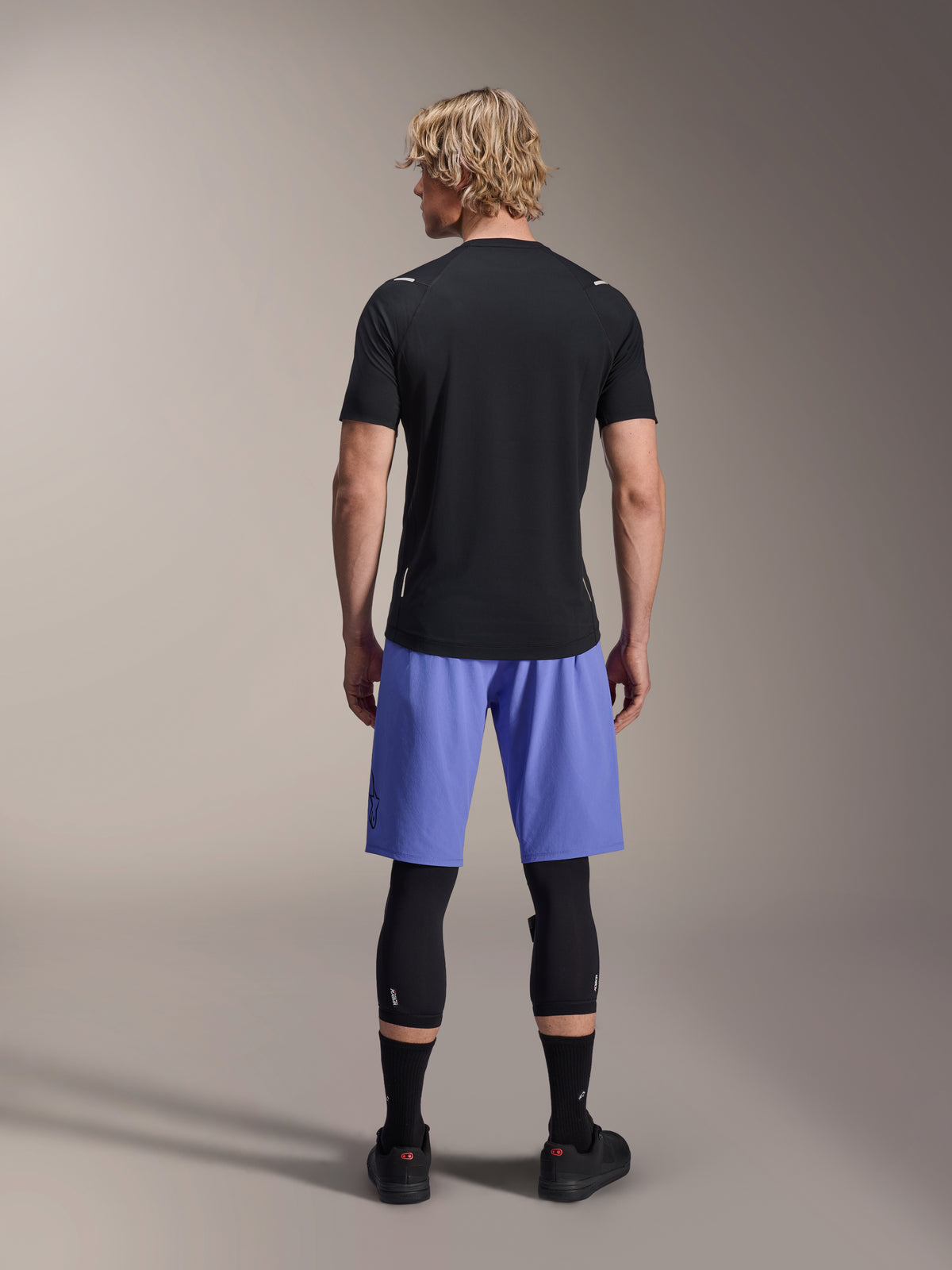 Rider uomo che indossa i pantaloncini Alpinestars A-Aria Lite, pantaloncini da MTB, Baja Blue, vista posteriore, abbinati a una maglia nera a maniche corte, ginocchiere nere e scarpe da ciclismo nere, abbigliamento per mountain bike ad alte prestazioni