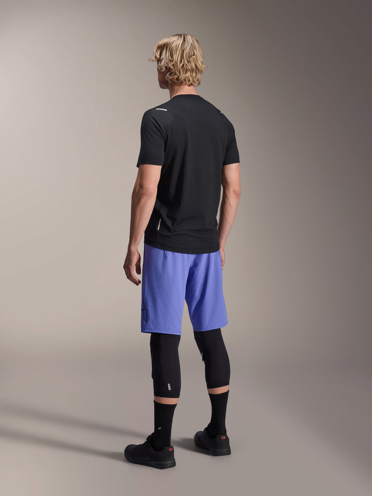 Alpinestars A-Aria Switch Jersey, maglia MTB a maniche corte, nero, vista posteriore su modello con pantaloncini blu e ginocchiere nere, abbigliamento tecnico da ciclismo per trail riding