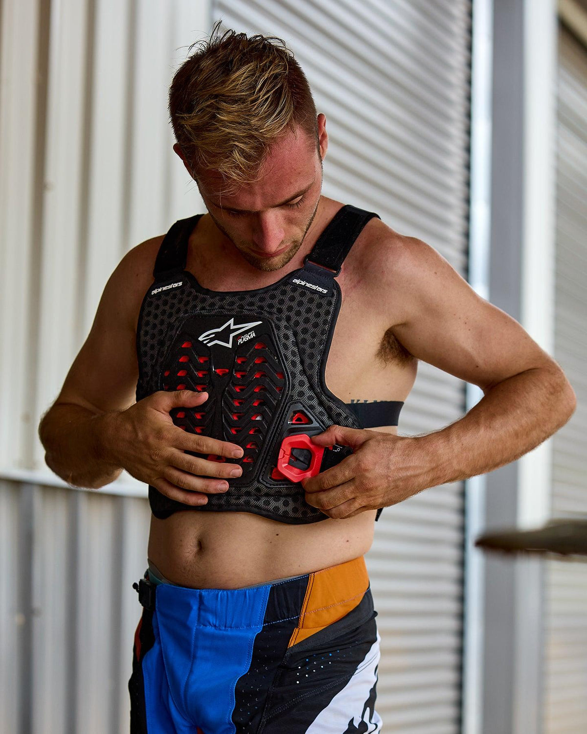 Uomo che indossa l'Alpinestars A-5 Plasma Chest Protector, nero con accenti rossi e logo bianco, con struttura a nido d'ape, abbinato a pantaloni da motocross blu e arancioni, in un contesto di preparazione per gare off-road