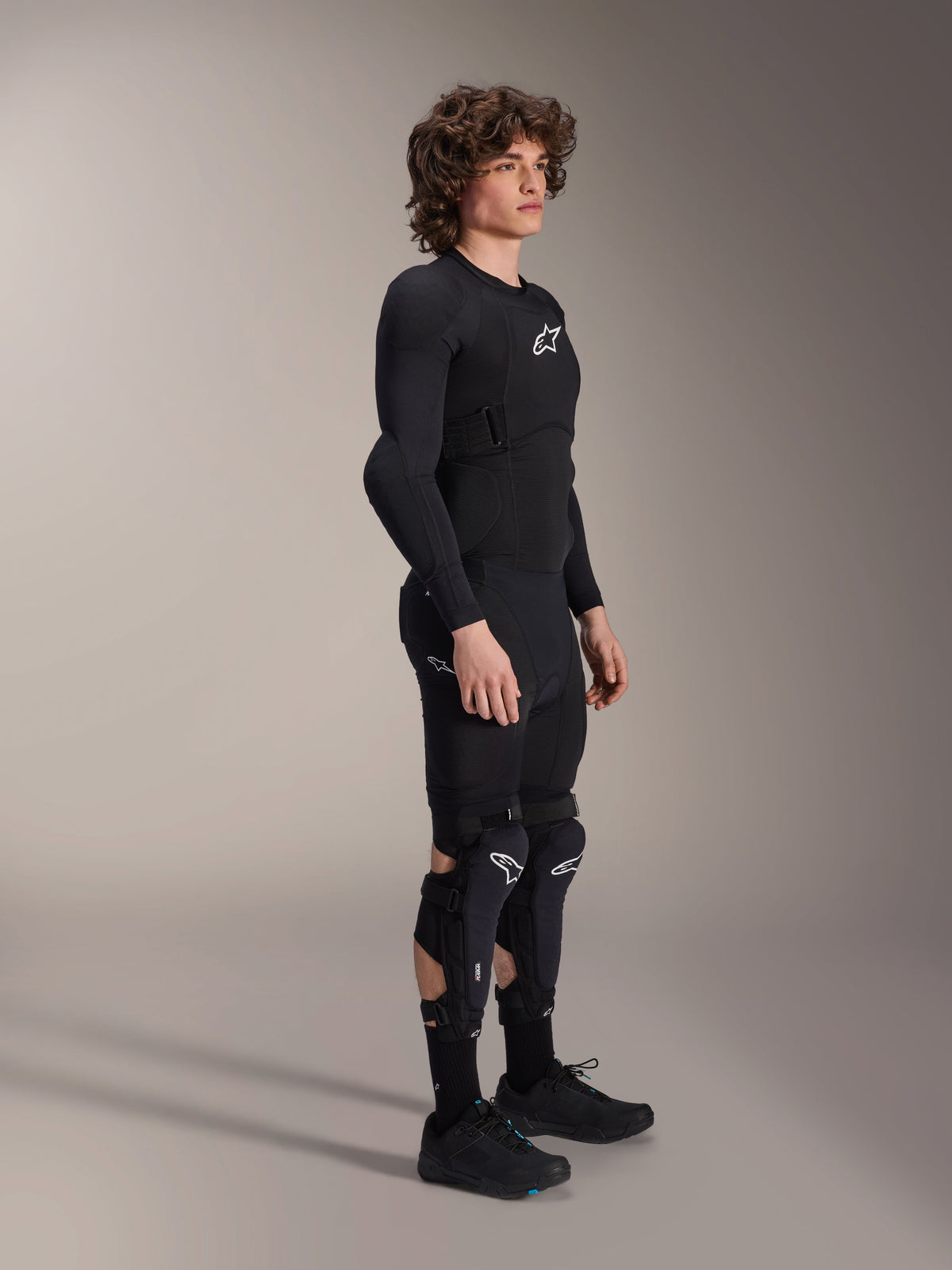 Giacca protettiva Alpinestars A-Impact Plasma - Maniche Lunghe, nera, indossata da un modello con equipaggiamento protettivo coordinato tra cui paraginocchia e pantaloncini, con loghi Astars bianchi, struttura in mesh traspirante e design aerodinamico per il ciclismo fuoristrada.