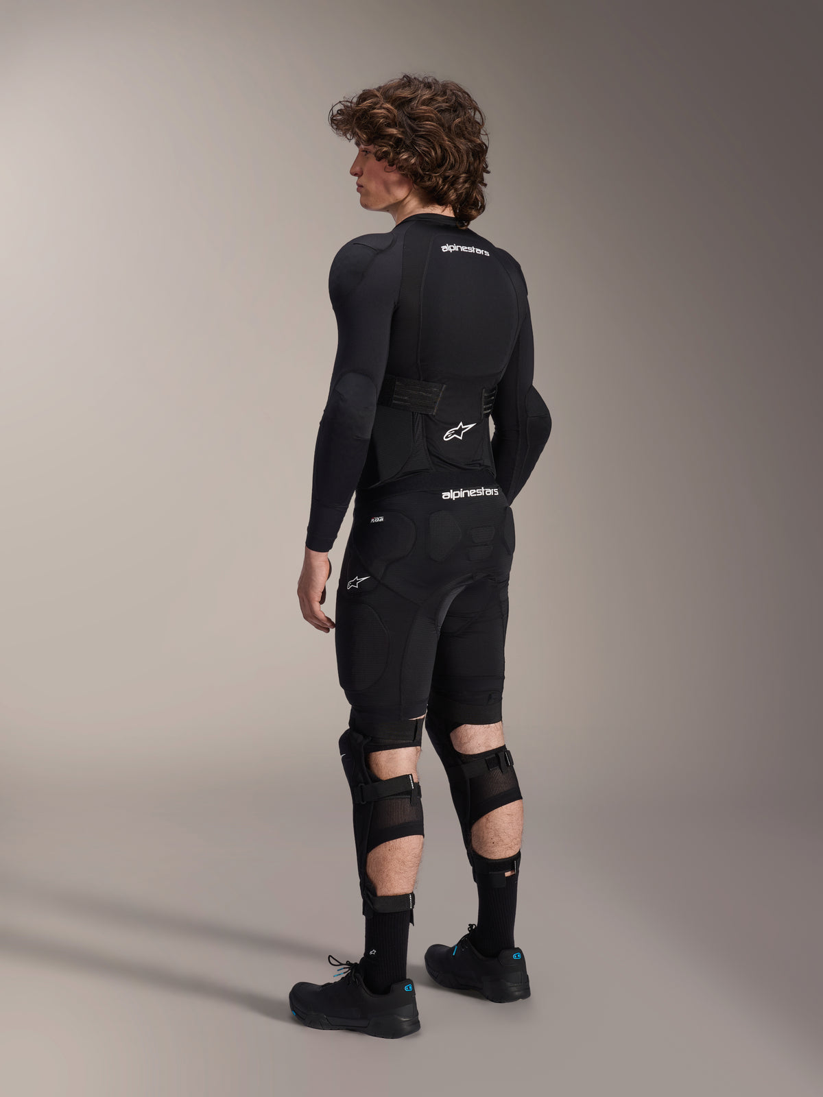 Modello che indossa protezioni ginocchio e stinco Alpinestars A-Impact Plasma Pro, vista dal retro, nero e bianco, con chiusura a triplo cinturino e pannelli in rete ventilata, indossate con un set completo di protezioni da mountain bike nero, base layer a maniche lunghe e pantaloncini imbottiti