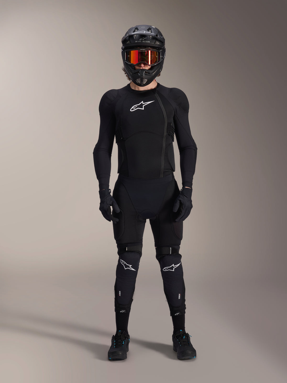 Ciclista di mountain bike con equipaggiamento protettivo completo, Giacca protettiva Alpinestars A-Impact Plasma in Nero e Bianco, con logo bianco sul petto e tessuto mesh, indossata con casco nero, maschera con lenti rosse e ginocchiere, set coordinato di protezioni MTB ad alte prestazioni.
