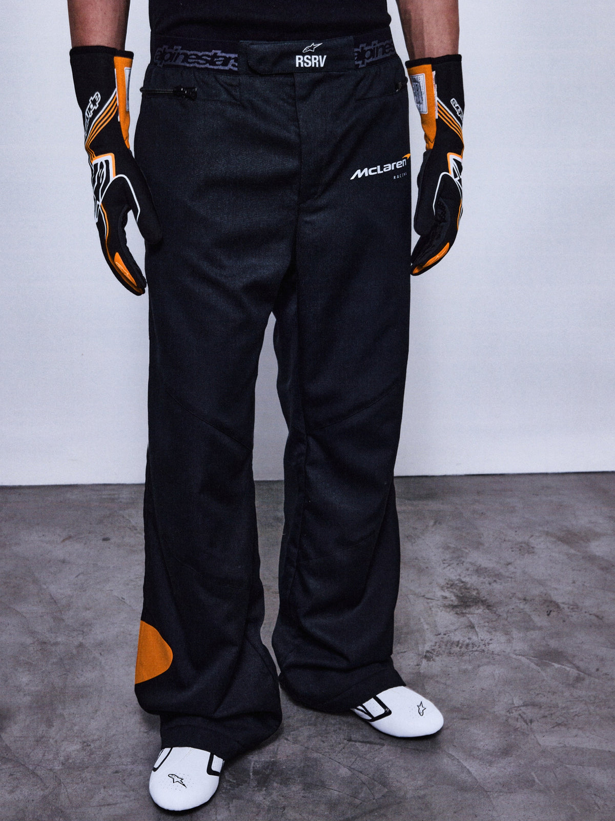 Alpinestars RSRV Arena Suit Pants, pantaloni da corsa neri con loghi McLaren e Alpinestars, dettagli arancioni, indossati con guanti racing neri e arancioni e scarpe bianche con accenti neri