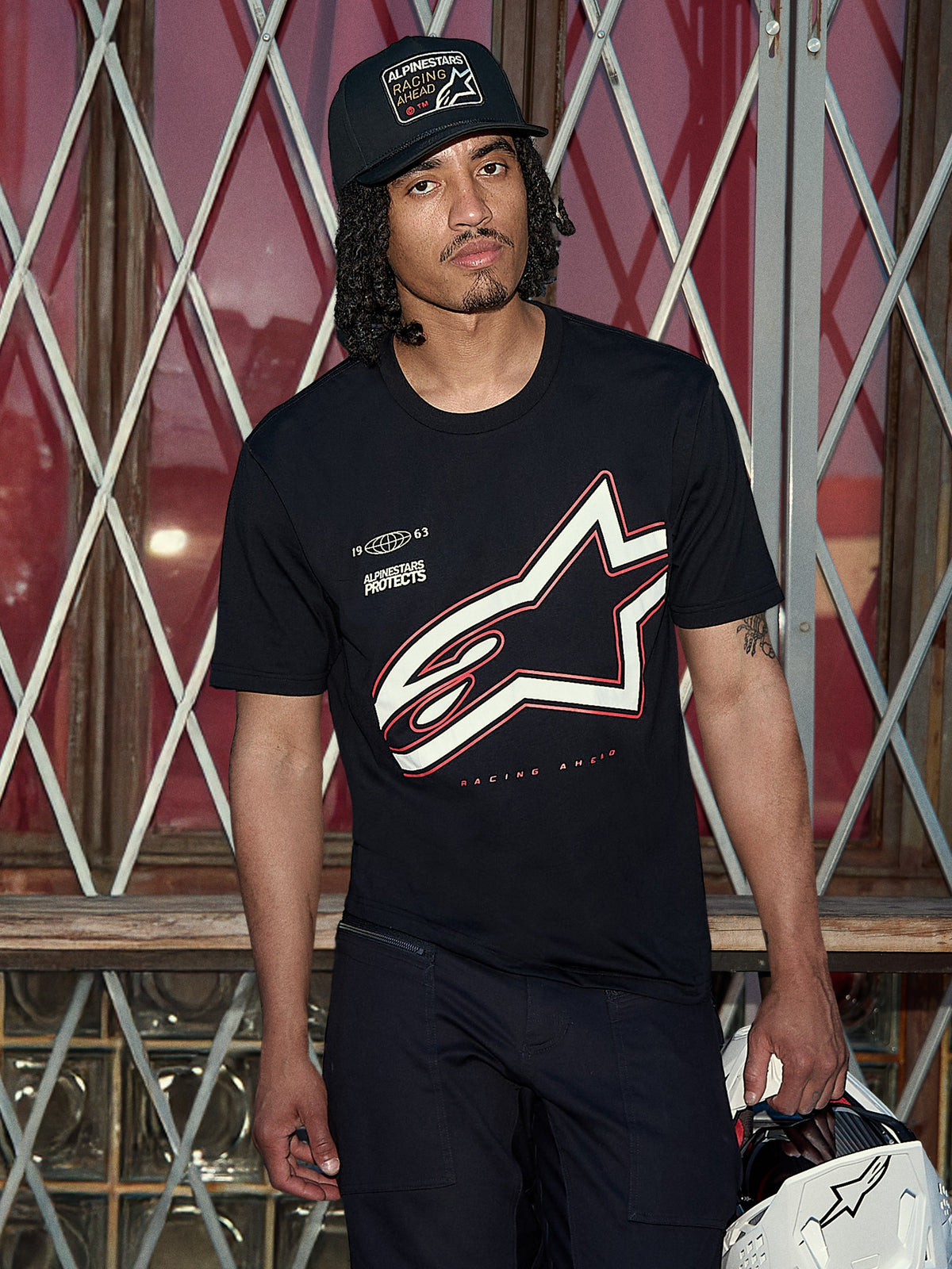 Alpinestars Authority CSF Tee, t-shirt a maniche corte, nera, grande logo Astars profilato bianco e rosso con grafica 'Alpinestars Protects', scatto lifestyle con un uomo che indossa un cappellino trucker nero e tiene un casco bianco, abbigliamento streetwear casual motorsport